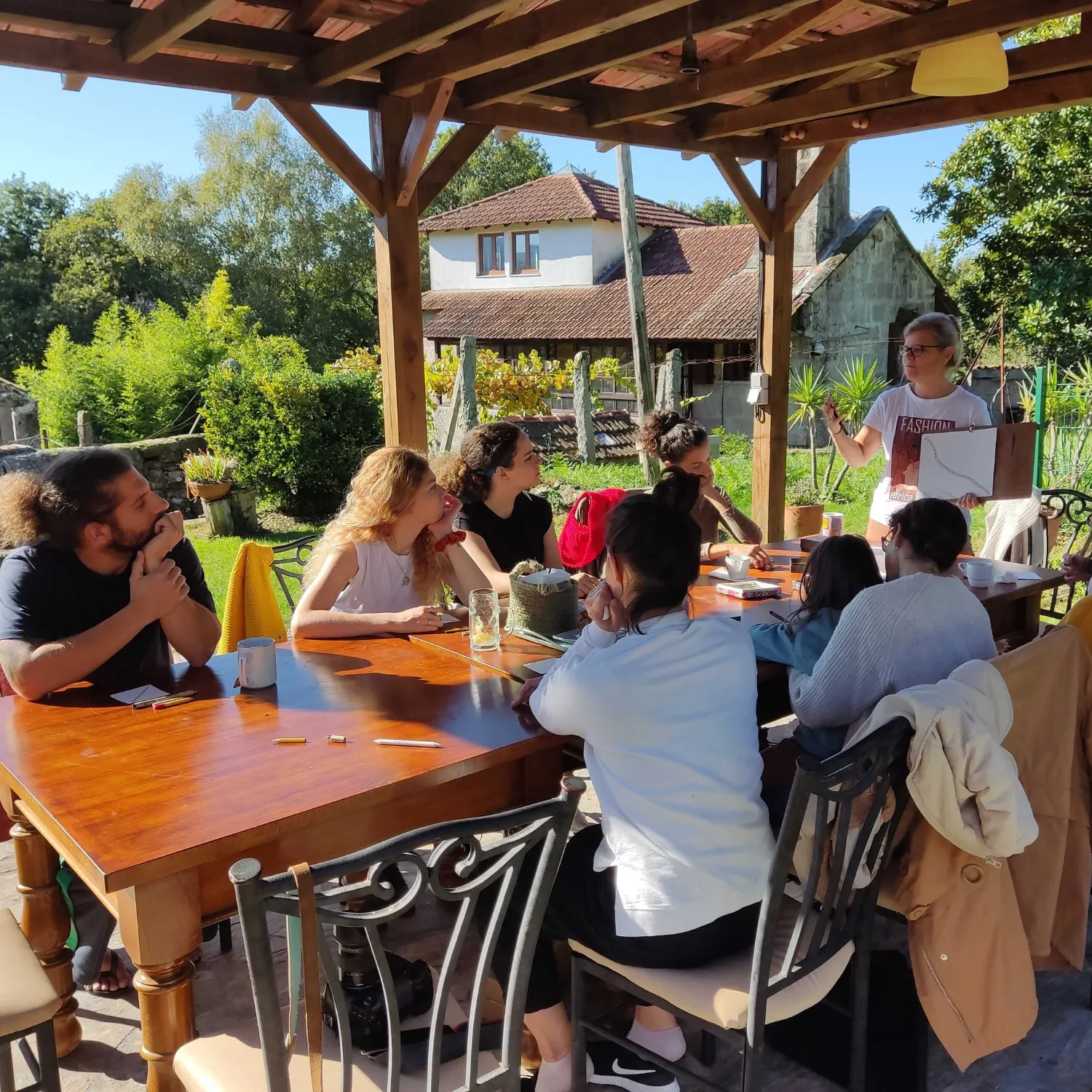 A woman holds up a drawing to a dozen people seated around a long wooden table under a pergola