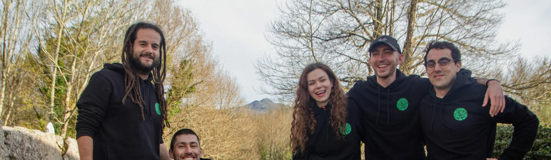 A wide crop of five smiling people in black hoodies with a small green logo, with bare trees behind them