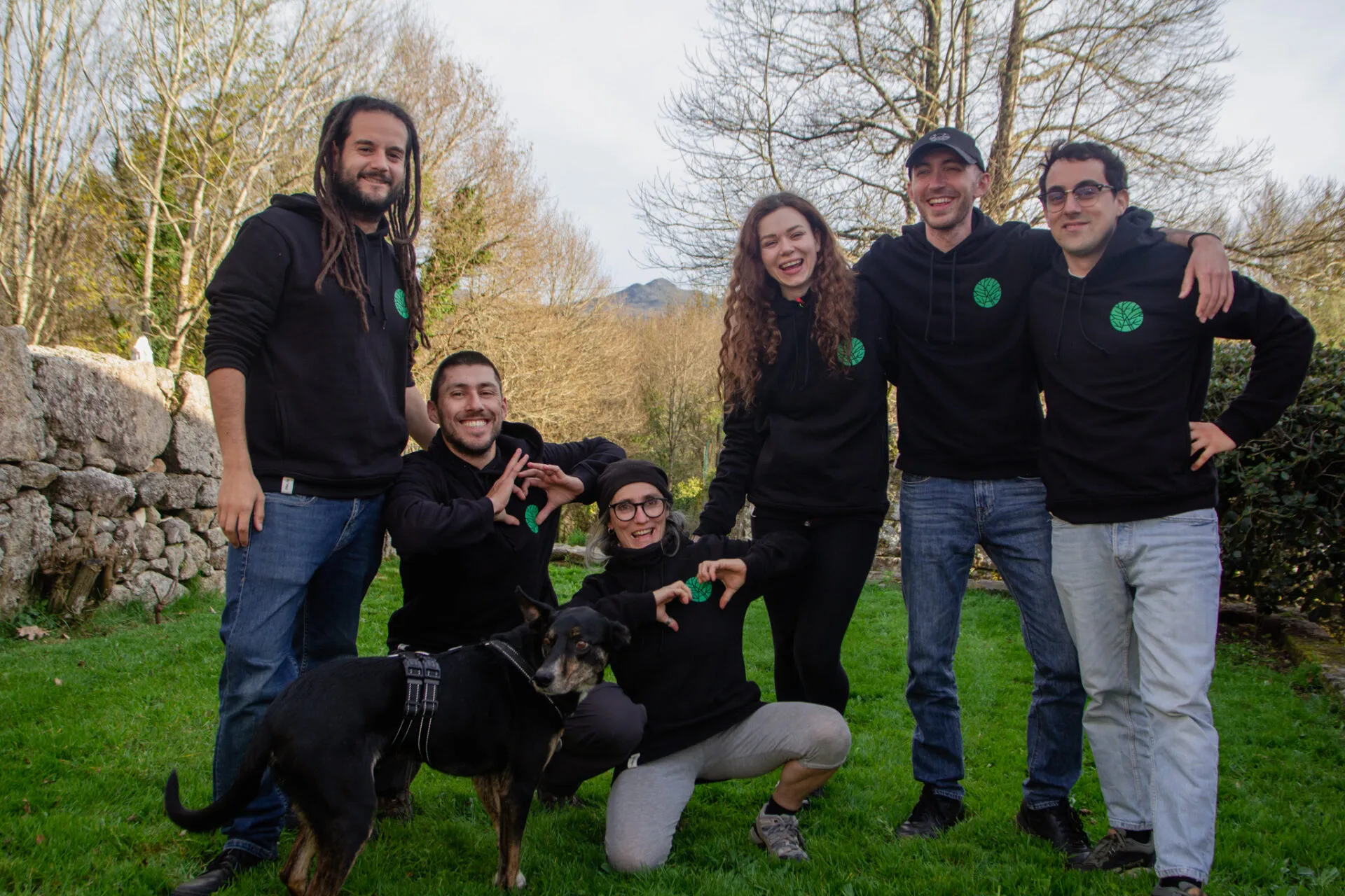 Six people in matching black hoodies with a green tree logo posing on grass with a black dog