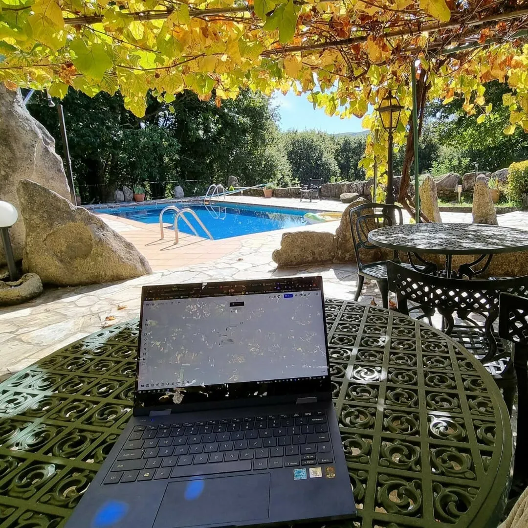 Open laptop on a green metal garden table under a vine pergola, with a blue swimming pool behind