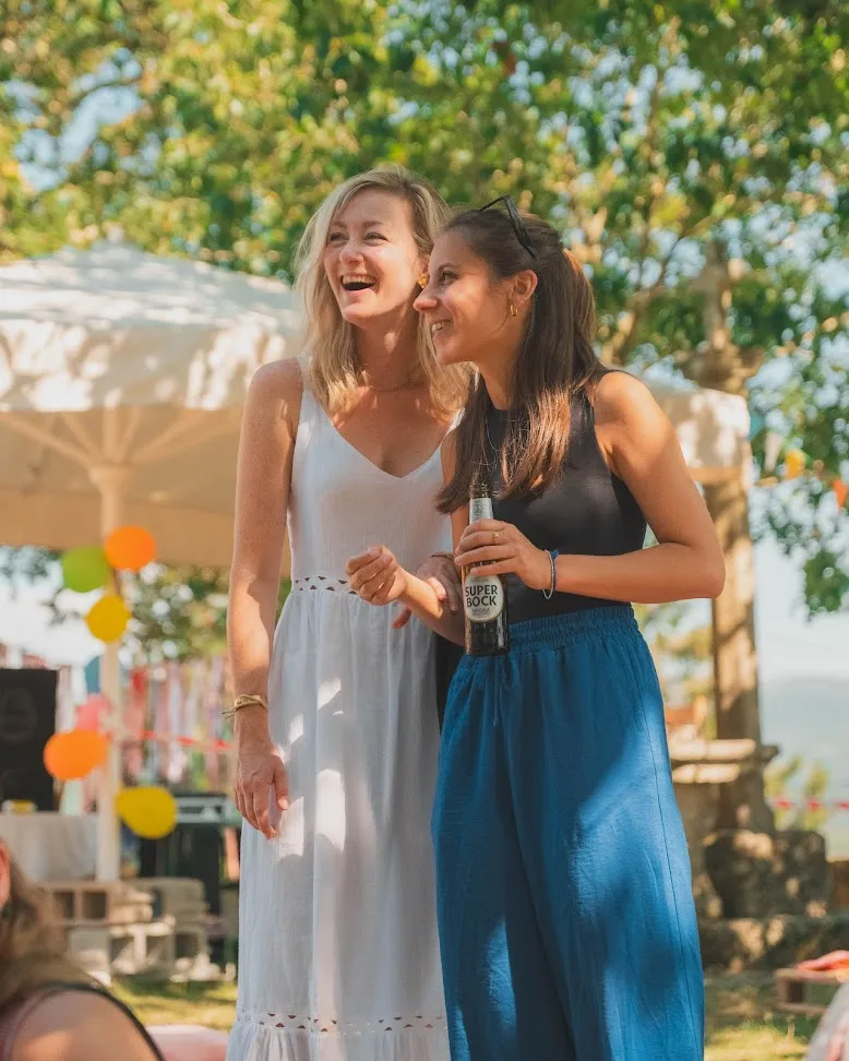 Two women laughing at an outdoor party, one in a white dress, the other holding a beer, bunting and a marquee behind