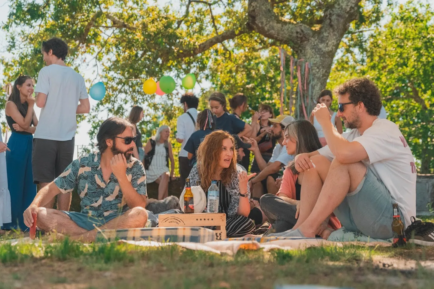 People sitting on picnic blankets under a tree with balloons, chatting in the sunshine
