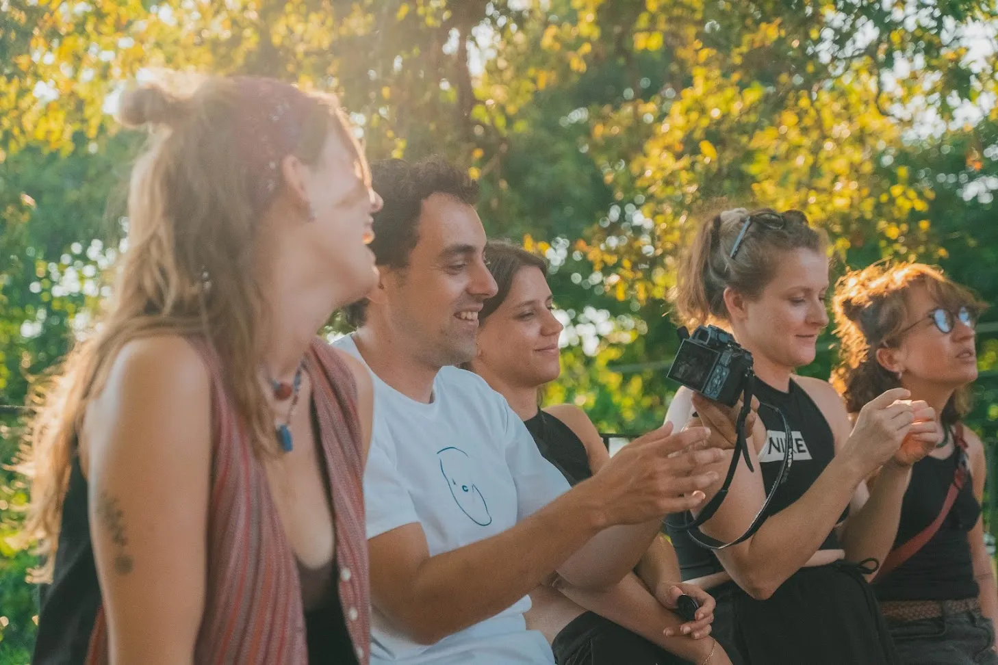 Five people sitting side by side in golden evening light, laughing while one holds up a camera