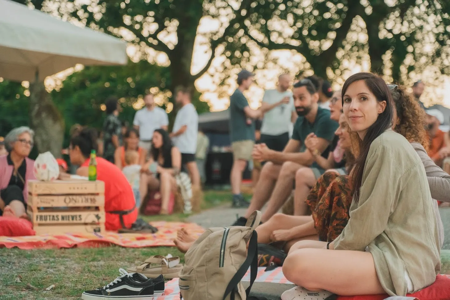 A woman looking at the camera among people sitting on blankets under the trees at a dusk gathering