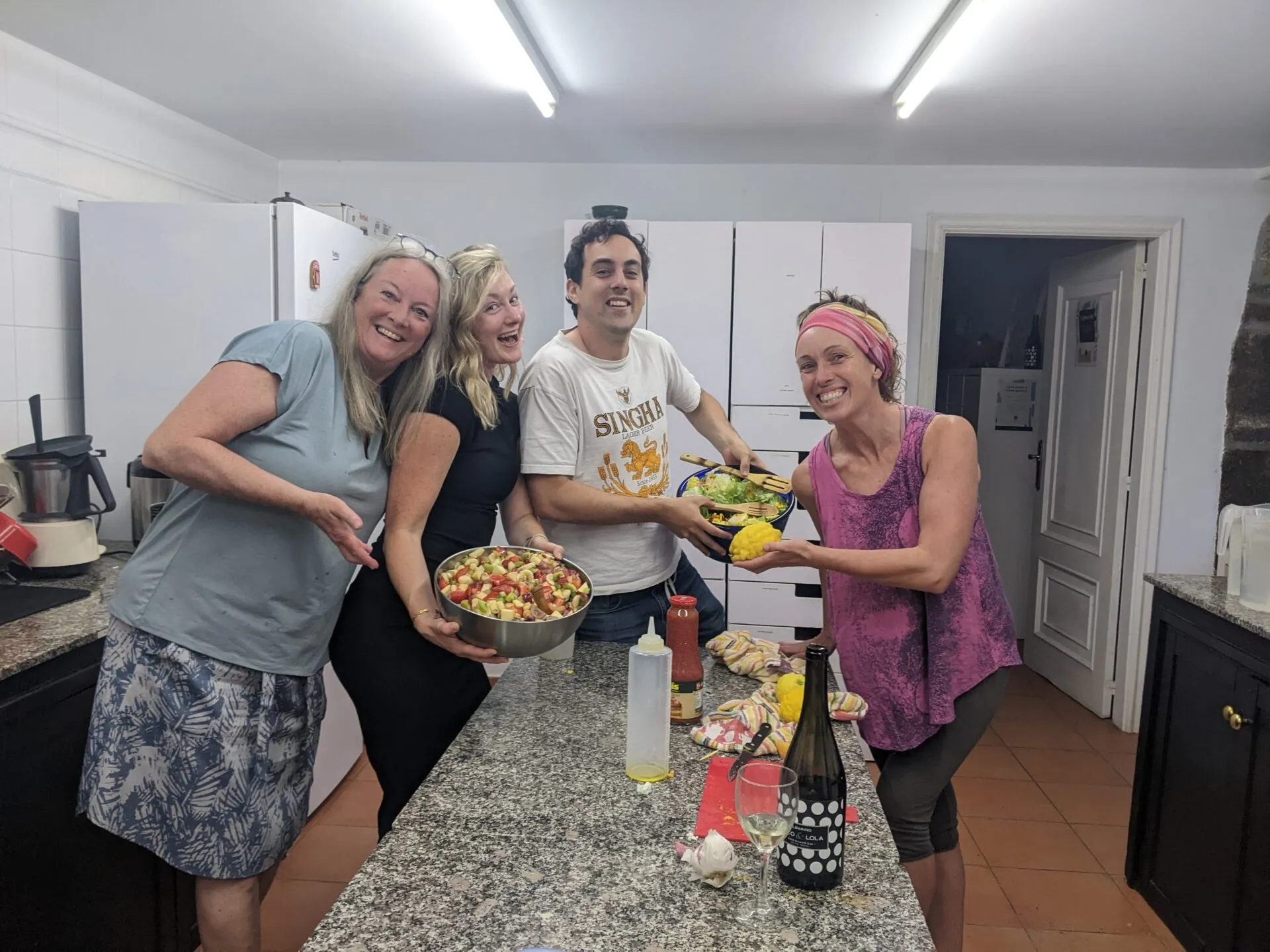 Four people laughing in a shared kitchen, holding big bowls of salad and chopped fruit