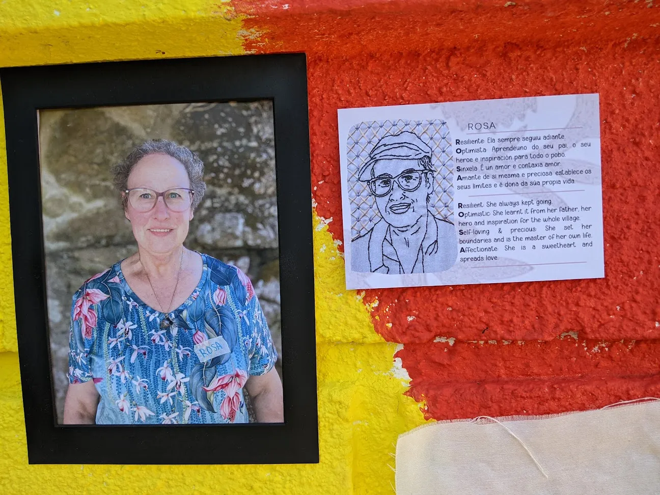 Framed portrait of a village woman beside an embroidered drawing and an acrostic text about Rosa, on a red and yellow wall