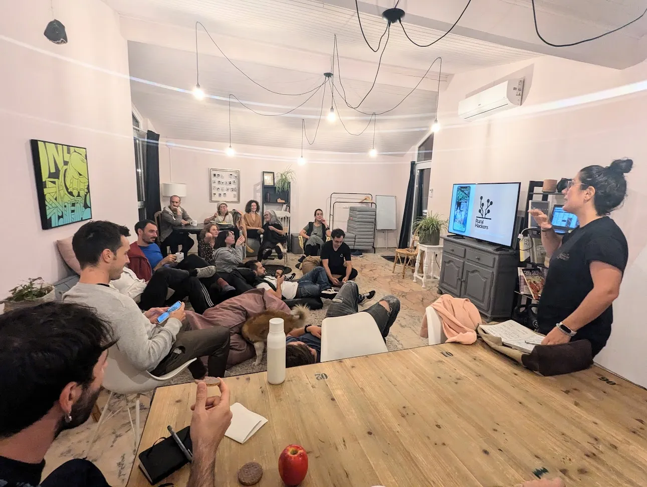 A woman presenting beside a screen showing the Rural Hackers logo, to a room of people seated and lying on the floor