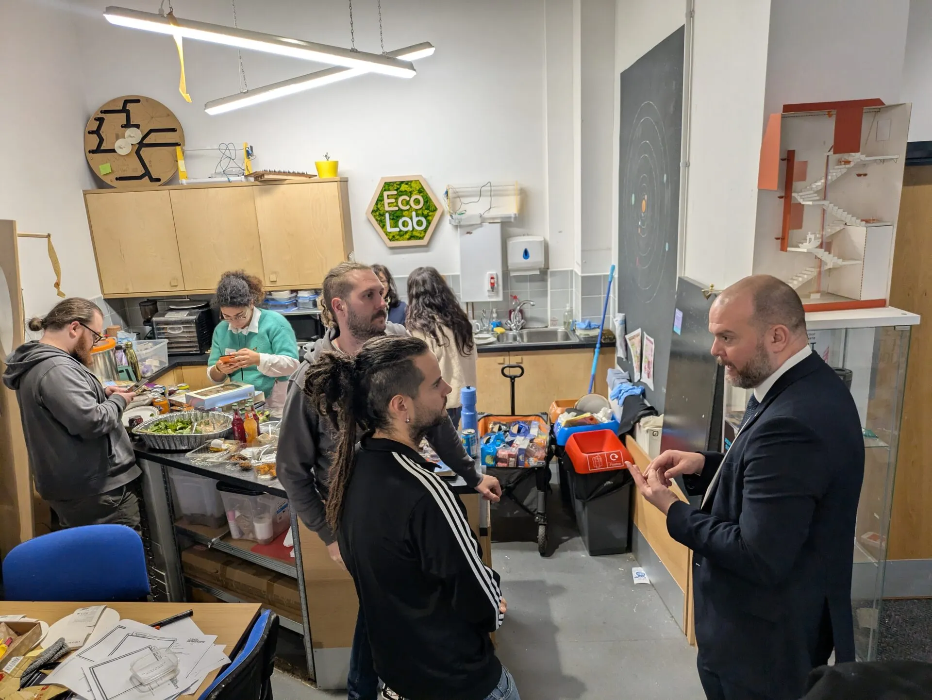 A man in a suit talking with visitors in the kitchen of the Eco Lab makerspace, food laid out on a trolley