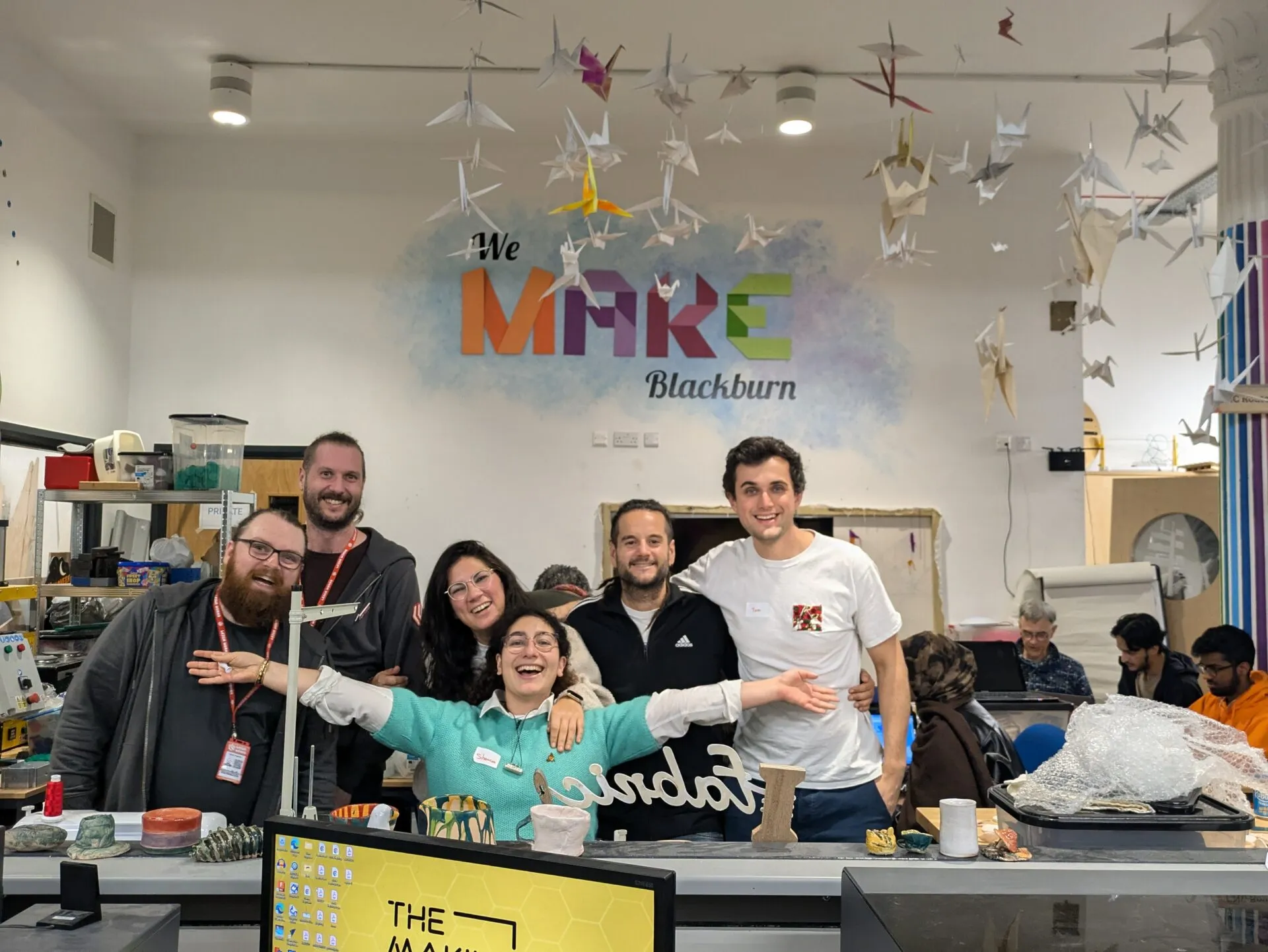 Six people laughing together in a makerspace beneath a wall logo reading ‘We Make Blackburn’, paper cranes hanging above