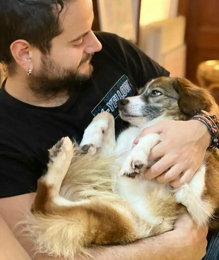 Bearded man in a black T-shirt cradling a small tan and white dog belly-up in his arms