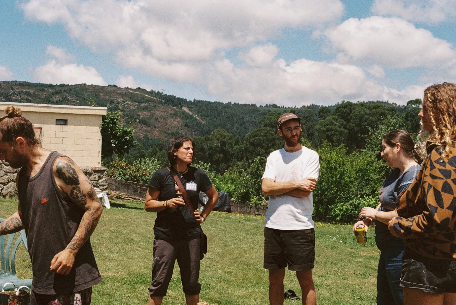 Five people standing and talking on a lawn, with wooded hills and a bright cloudy sky behind them