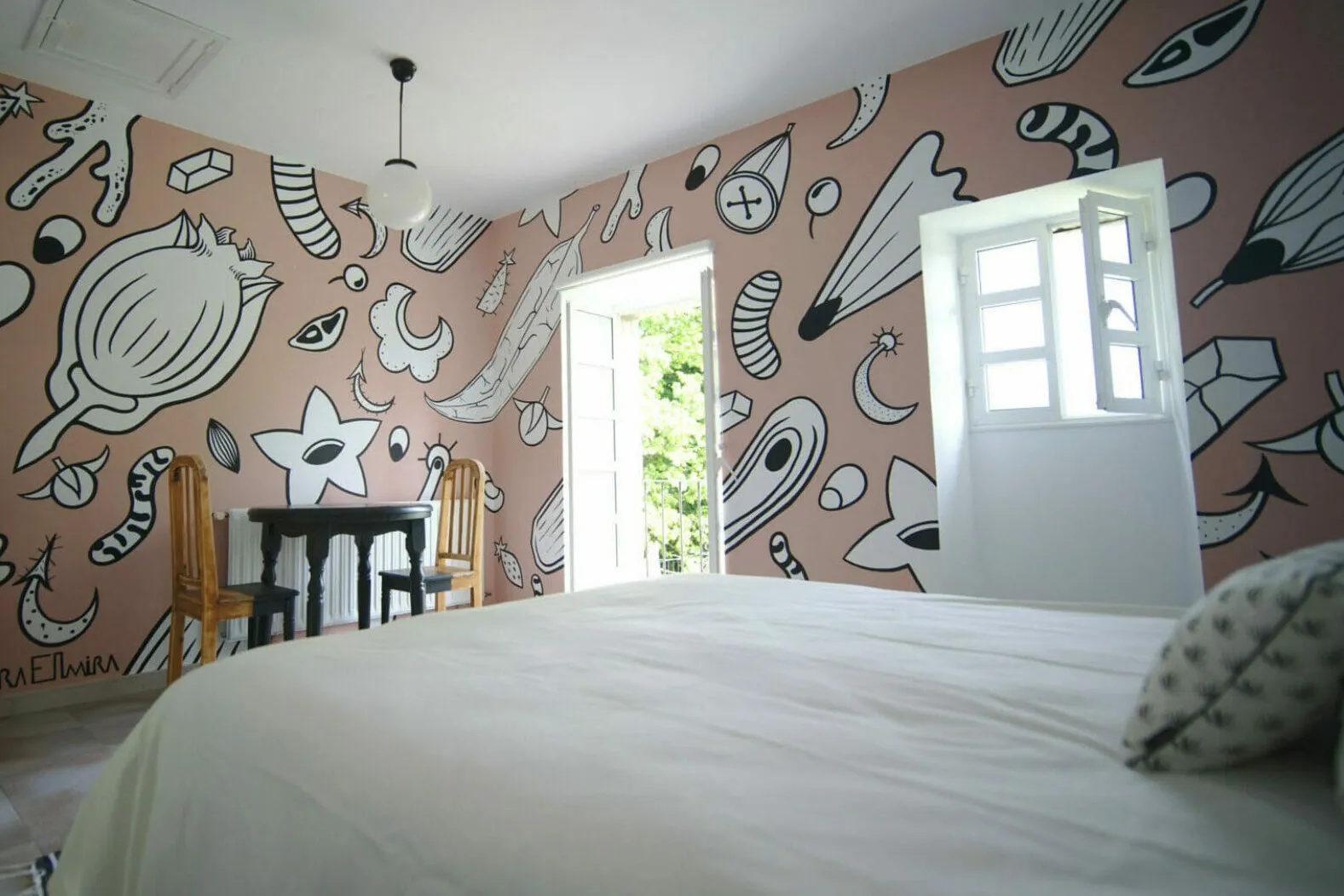 A bedroom with dusty pink walls covered in black and white drawings of seeds, shells and moons, and a door open onto the green outside