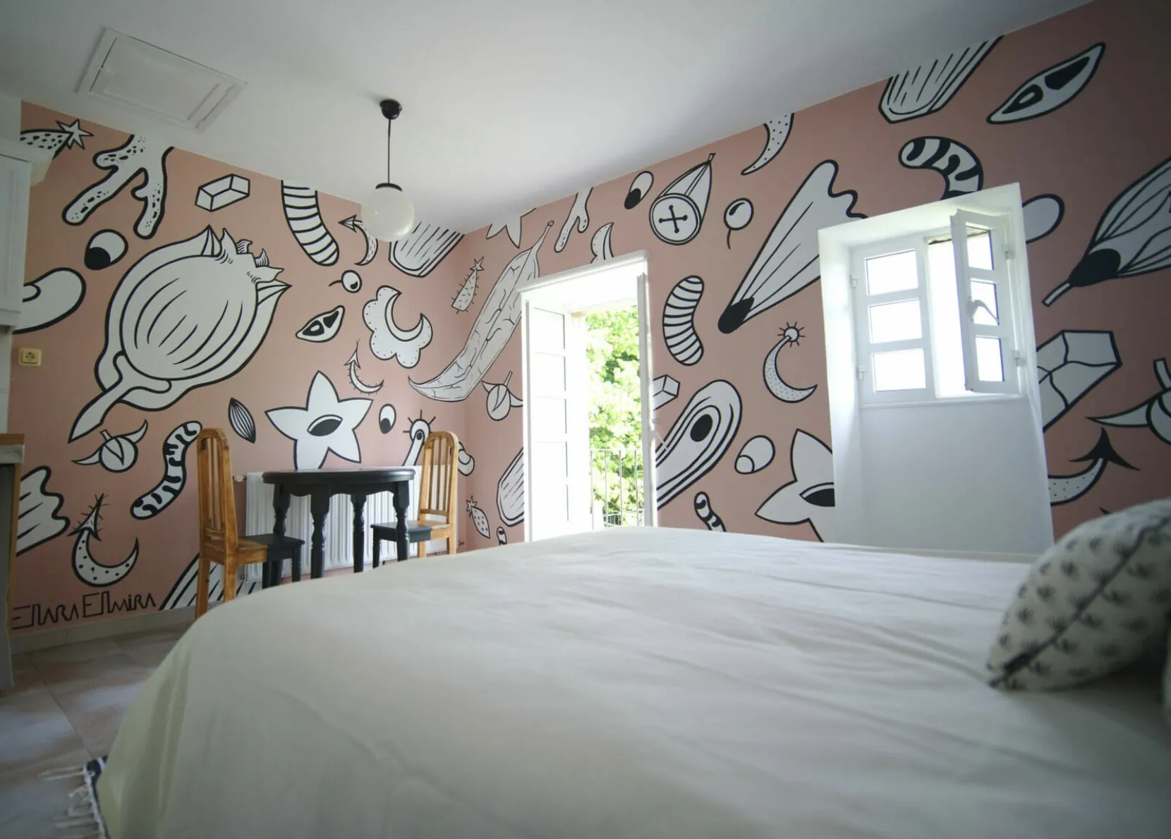A bedroom with a large white bed and walls painted with black-and-white doodles on a pink background, with open balcony doors