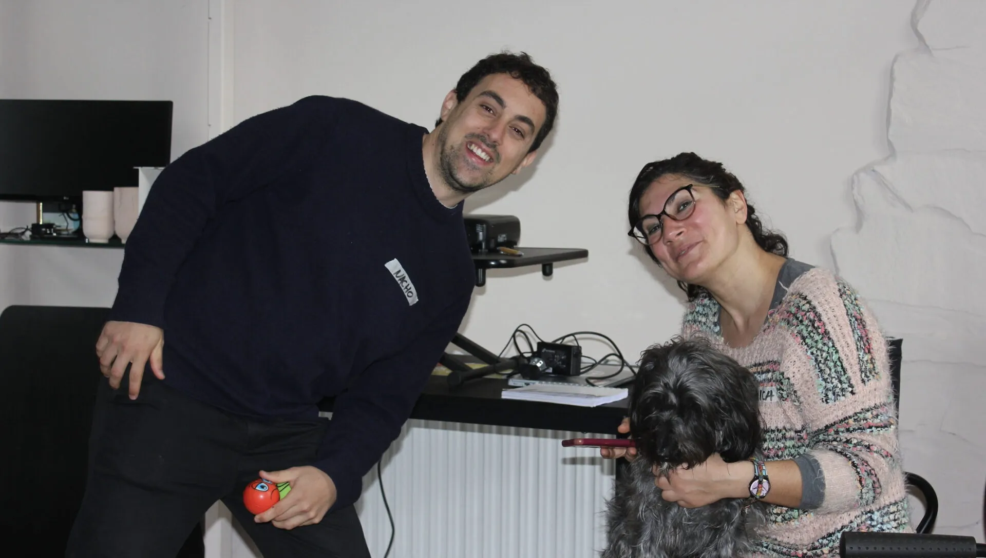 A grinning man in a navy jumper leaning beside a woman in glasses hugging a shaggy grey dog in an office