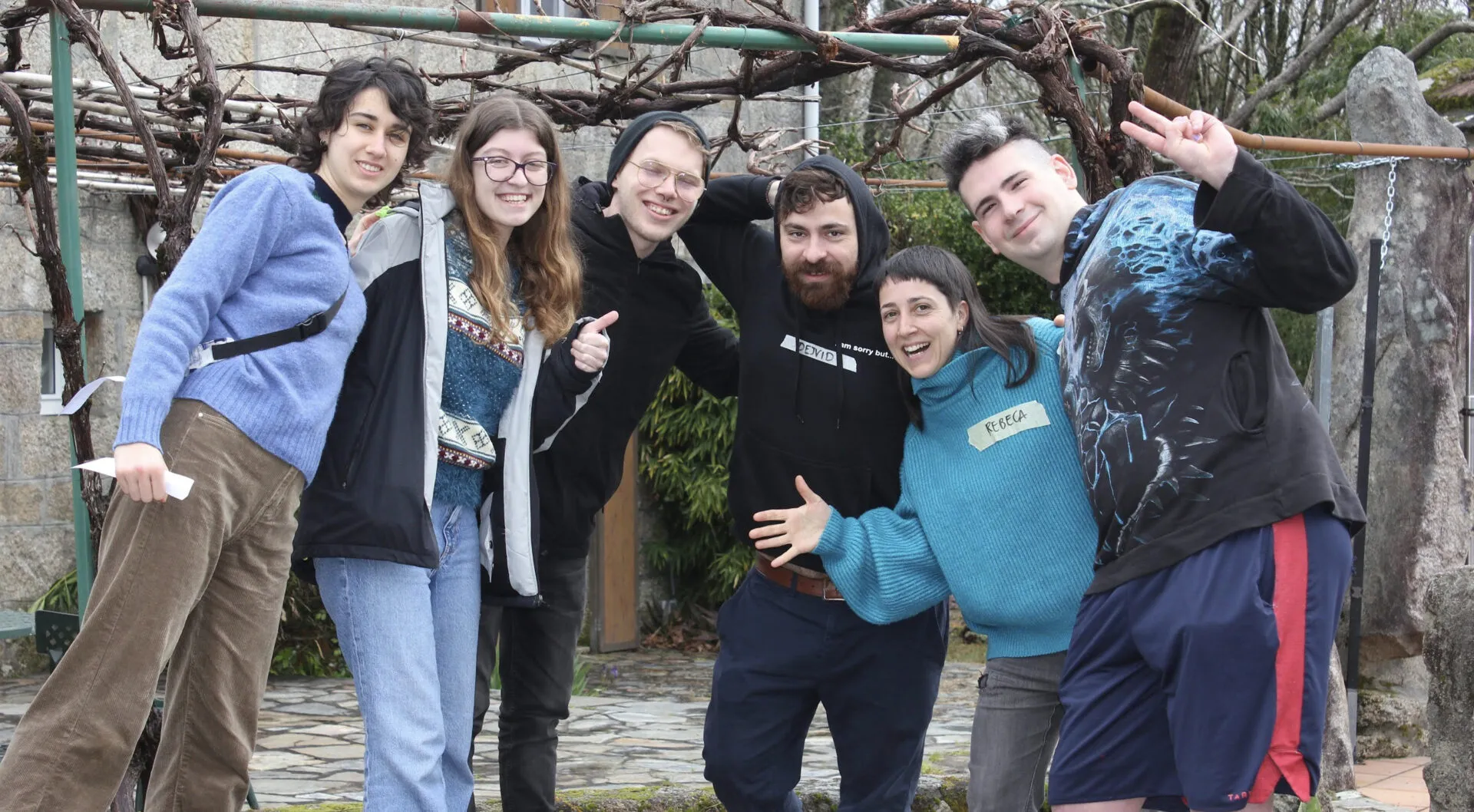 Six young people posing playfully arm in arm under a bare grapevine pergola, one wearing a paper name tag reading Rebeca