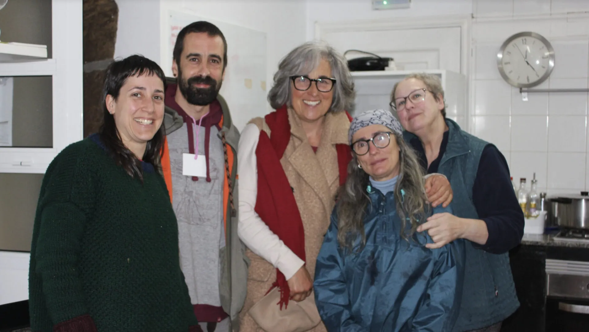 Five people smiling arm in arm in a white-tiled communal kitchen with a wall clock behind them