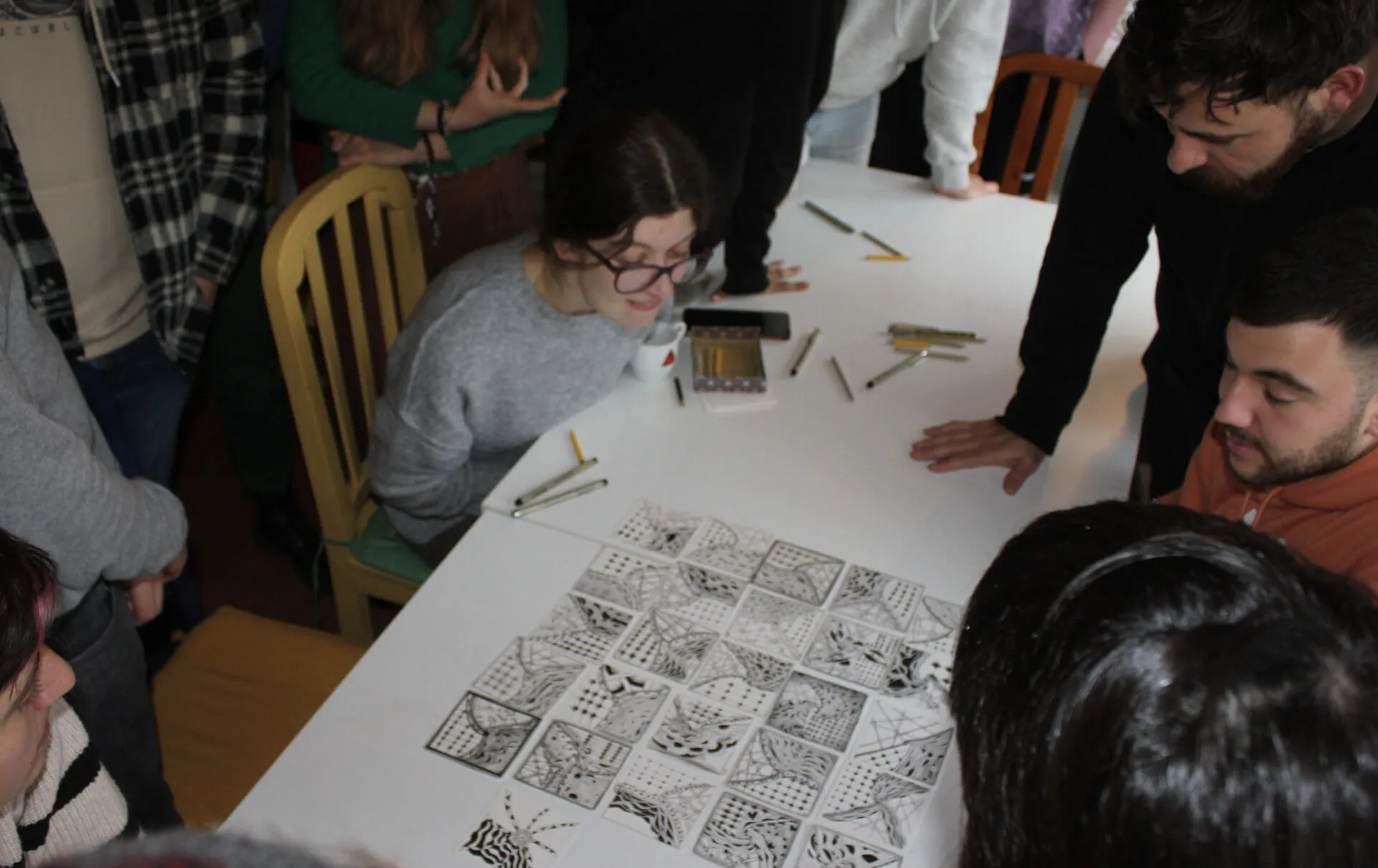 People leaning over a white table covered with hand-drawn black-and-white patterned tiles and scattered pens