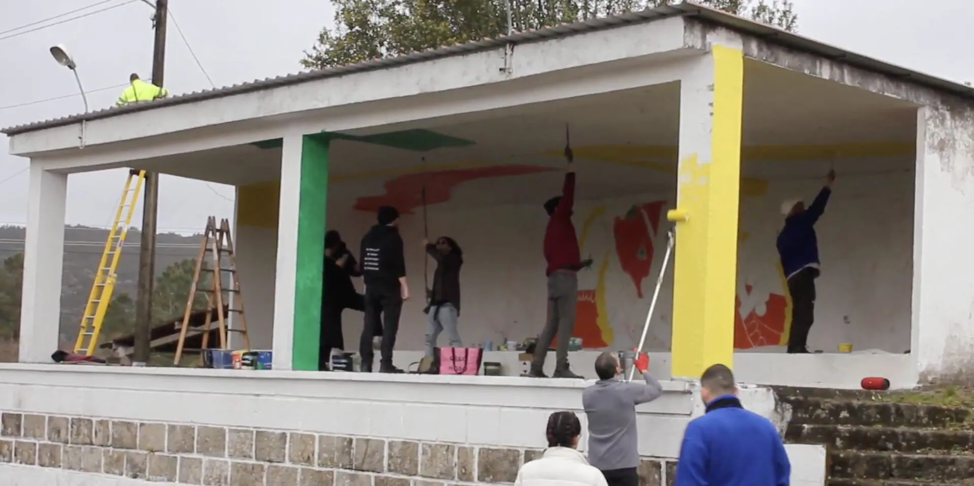 Six people on a village bandstand painting a large colourful mural, with ladders and paint pots around