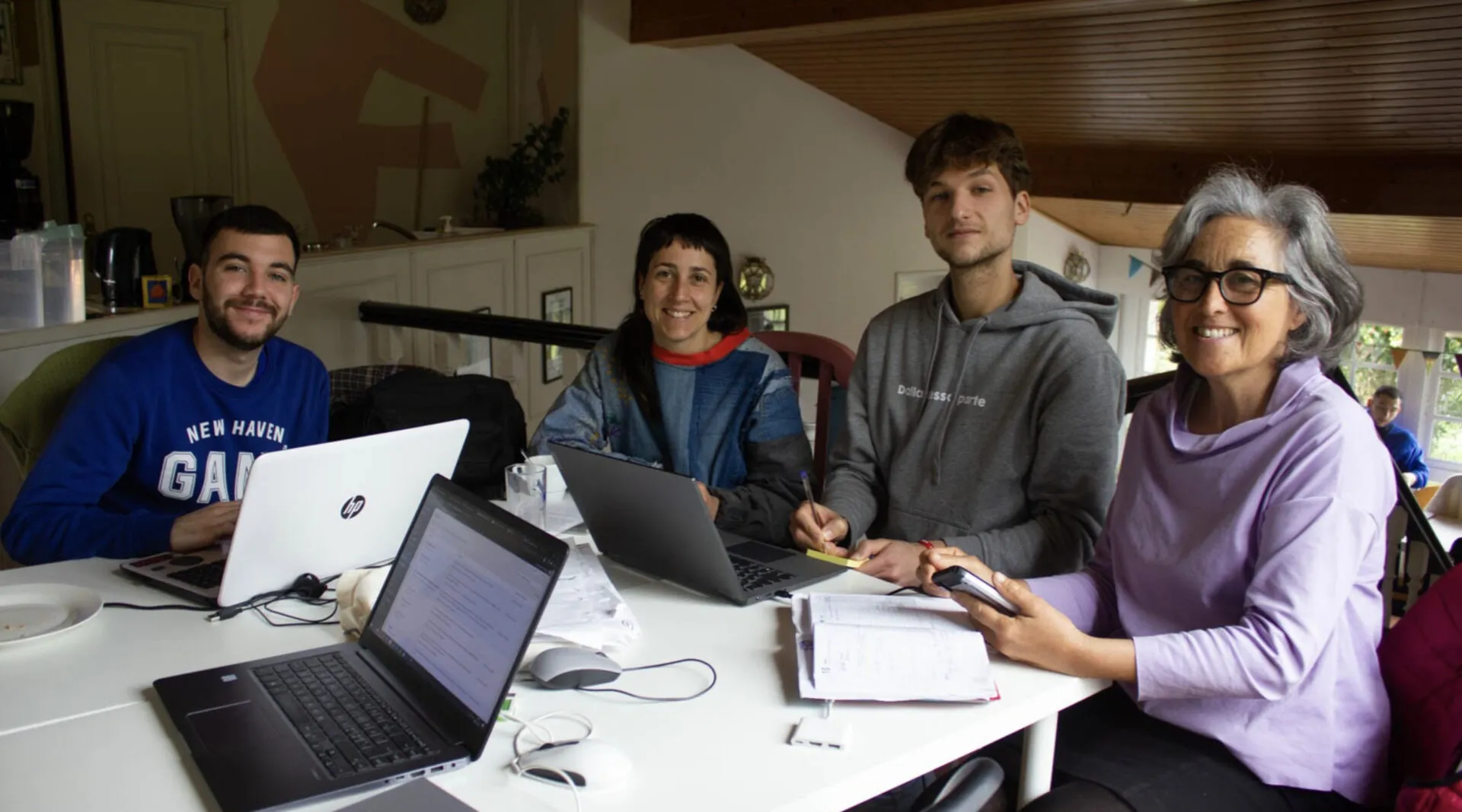 Four people smiling at a white table covered with laptops and papers in a bright room with a wooden ceiling