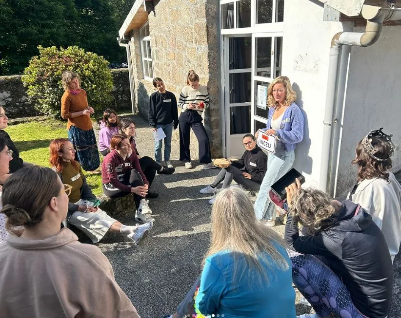 Group sitting in a circle outside a stone building while a woman standing against the wall holds up a card reading Anceu