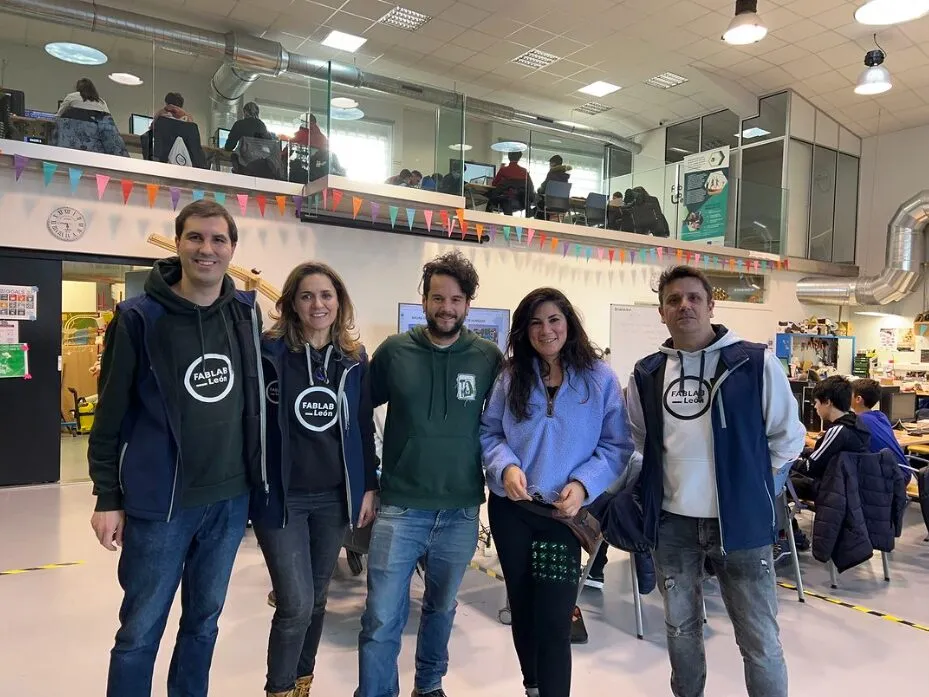 Five people posing in a fab lab, three of them in FABLAB León hoodies, with bunting and a mezzanine of computer desks behind