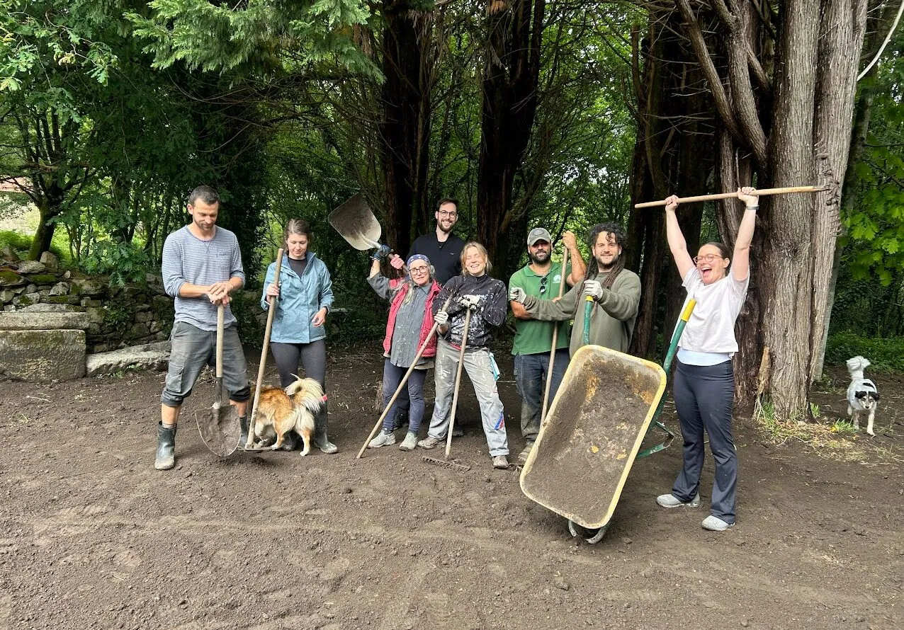 Eight people posing with shovels, rakes and a raised wheelbarrow on freshly turned earth, two dogs beside them