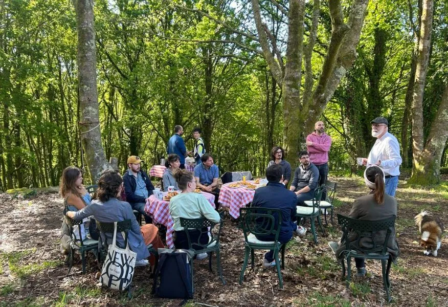 A dozen people seated in a circle of garden chairs in an oak wood, listening to a man standing beside tables laid with food