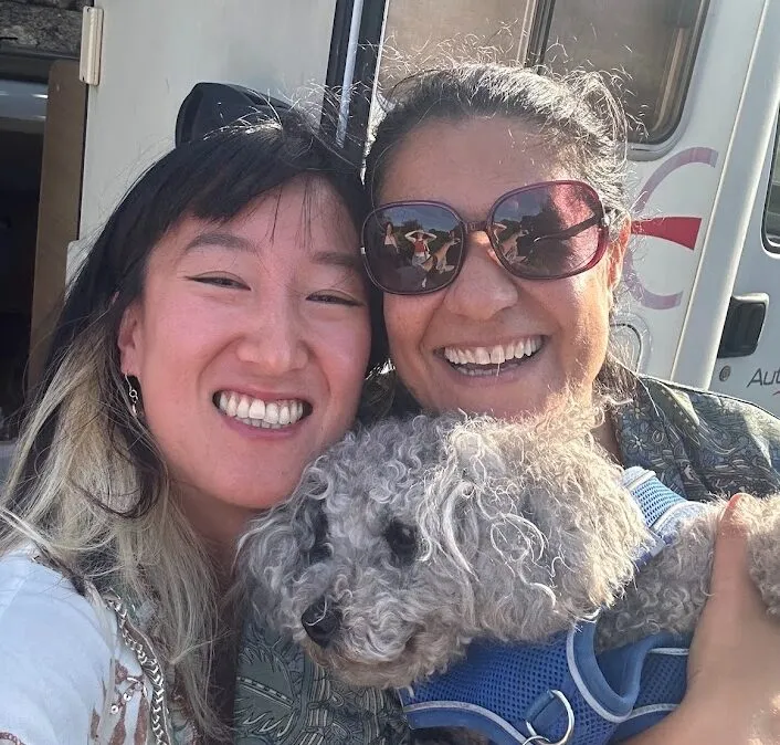 Selfie of two women cheek to cheek holding a small curly grey dog in front of a campervan