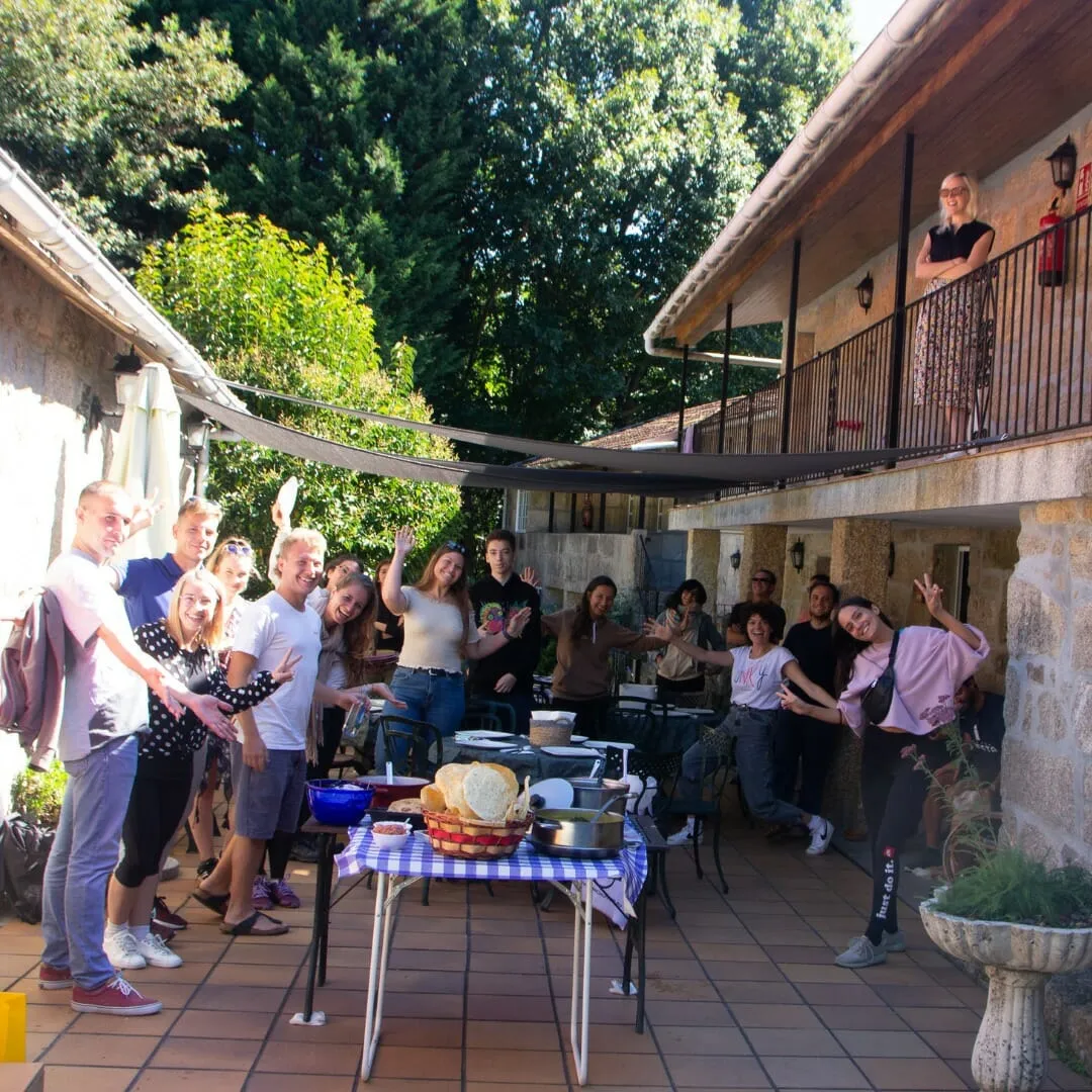 About fifteen people waving and posing on a stone-house terrace beside a table laid with bread and shared food