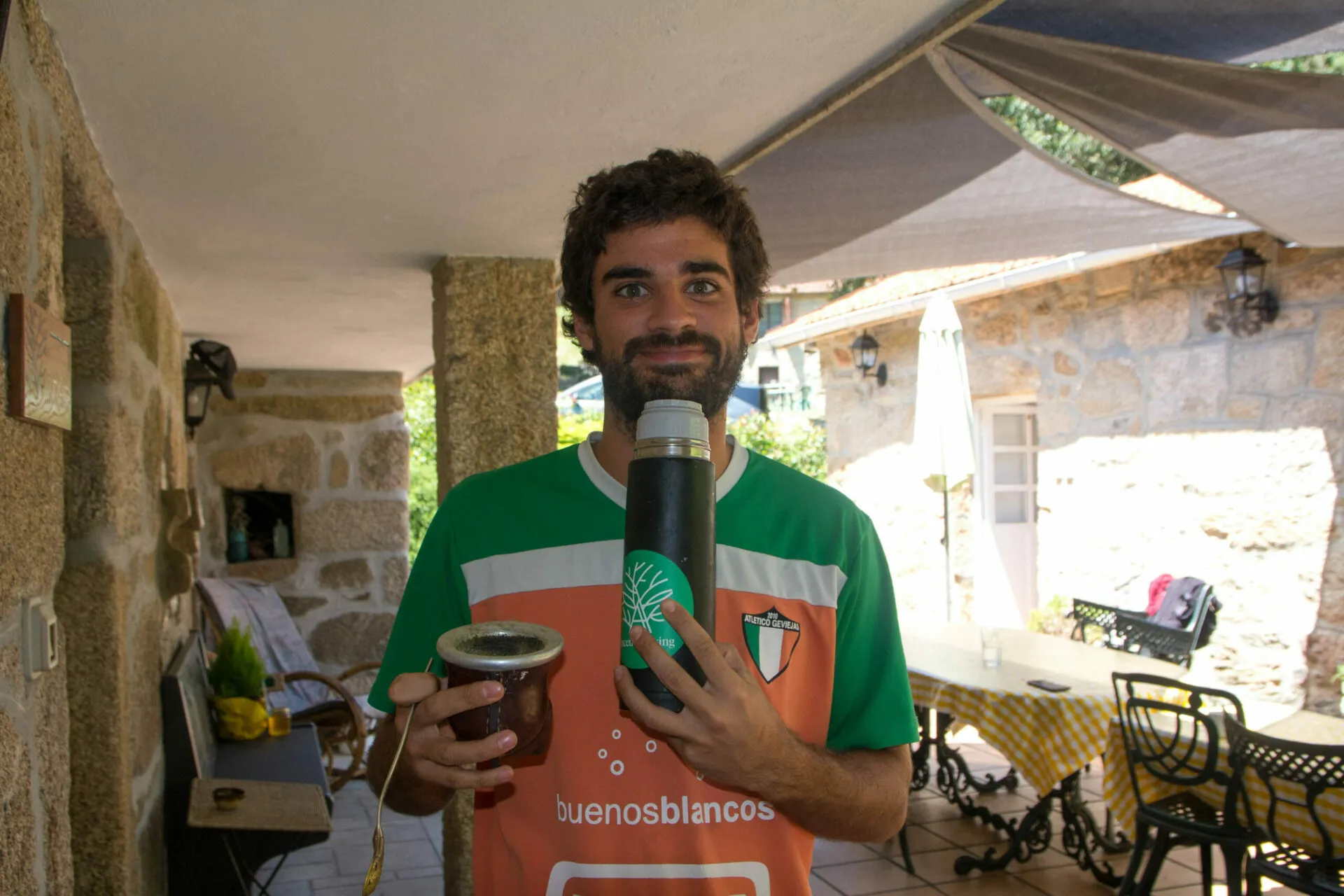 Young bearded man in an orange and green football shirt holding a mate gourd and a thermos on a stone terrace