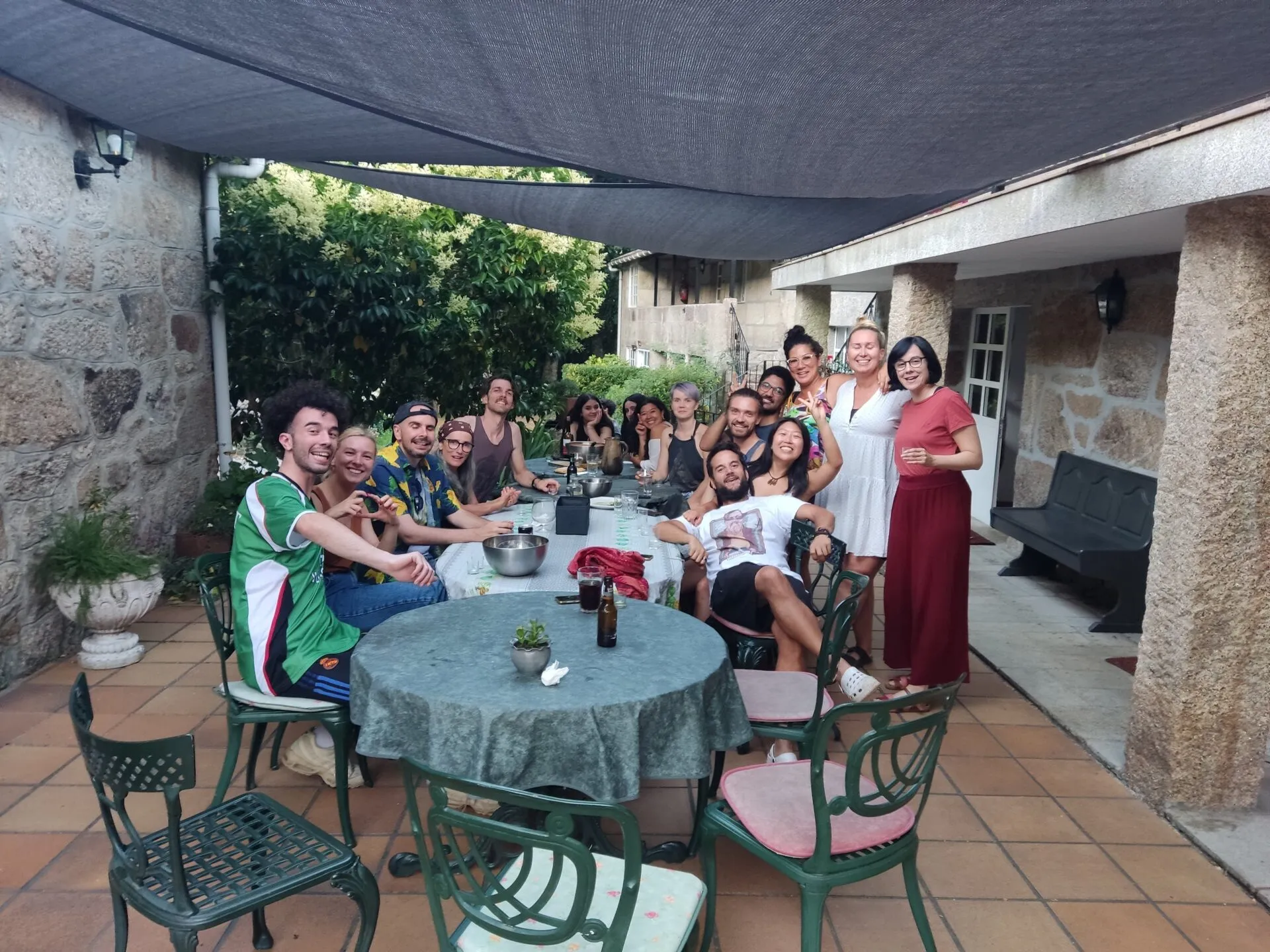 A long table of friends on a covered stone terrace, some smiling towards the camera, with a green tablecloth on the table in front