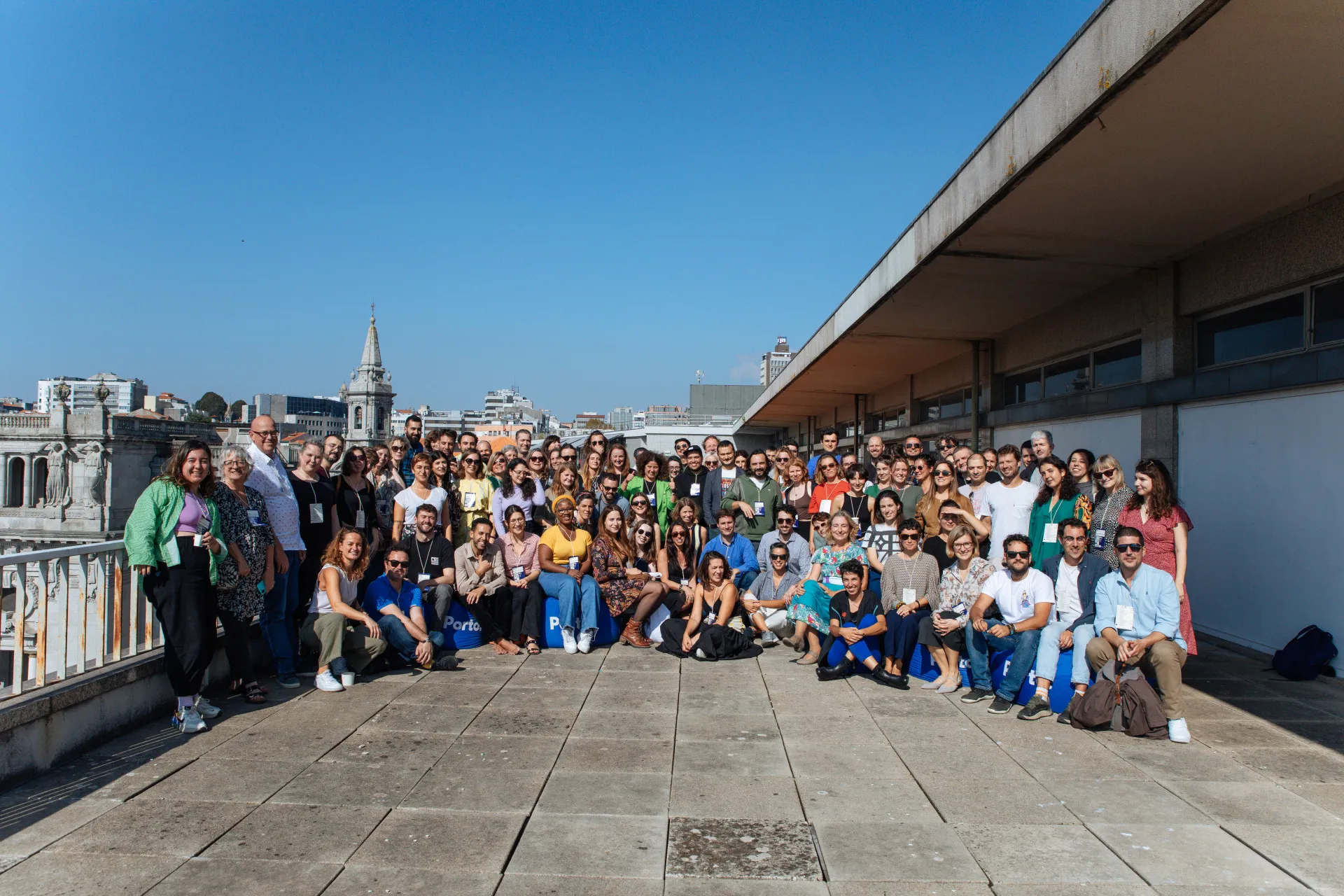 A crowd of more than a hundred people posing on a paved rooftop terrace under a blue sky, with city buildings behind