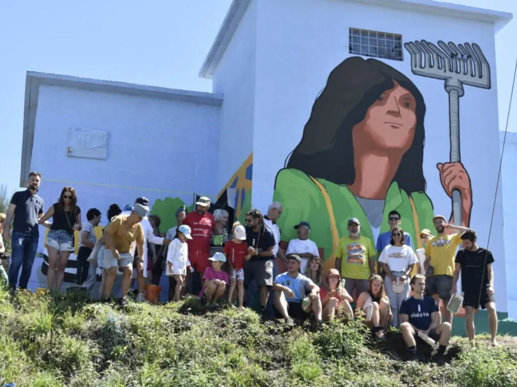 Villagers and colivers gathered below a mural of a woman holding a rake painted on a white building