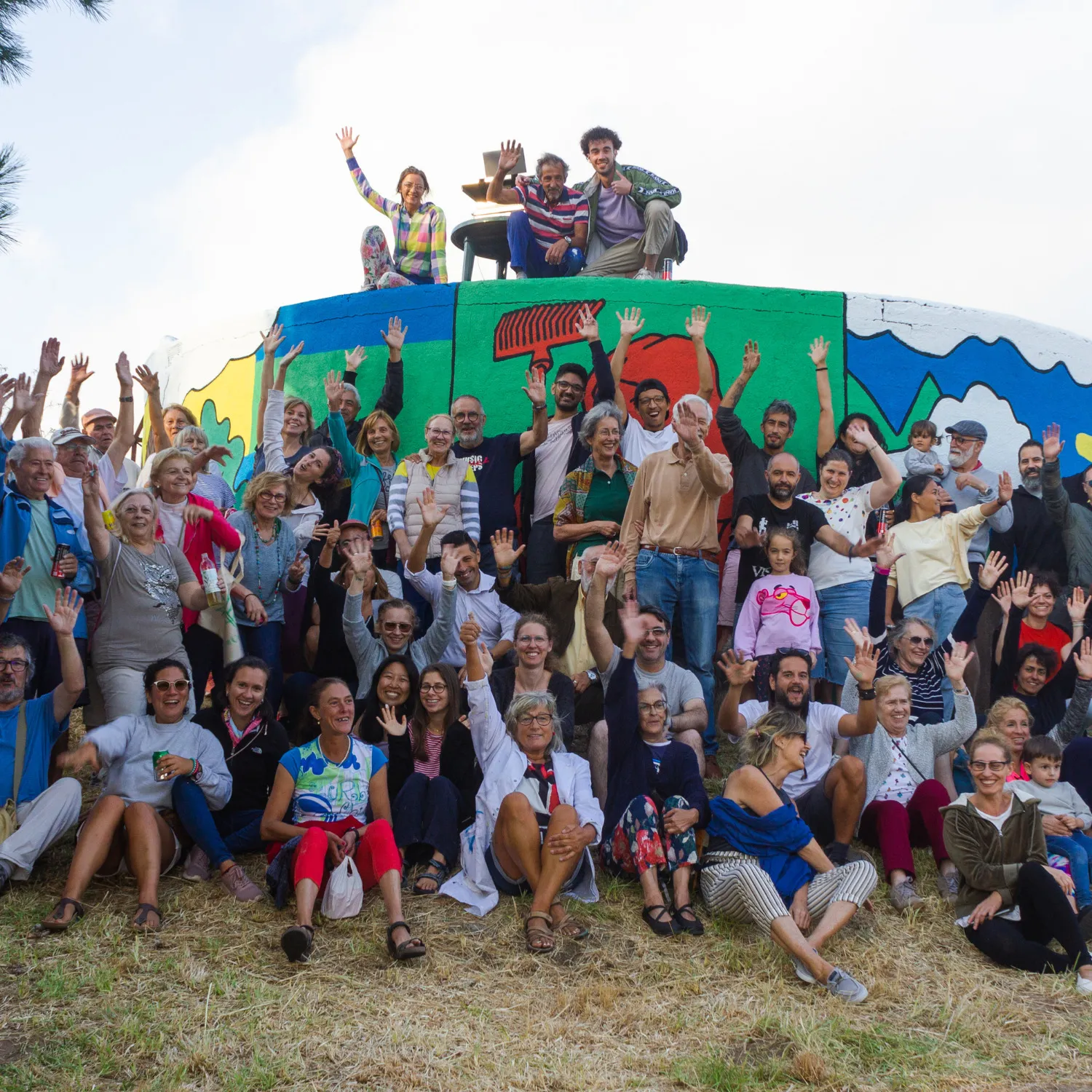 Large crowd of villagers and colivers waving in front of a water tank painted with a colourful mural, three people sitting on top