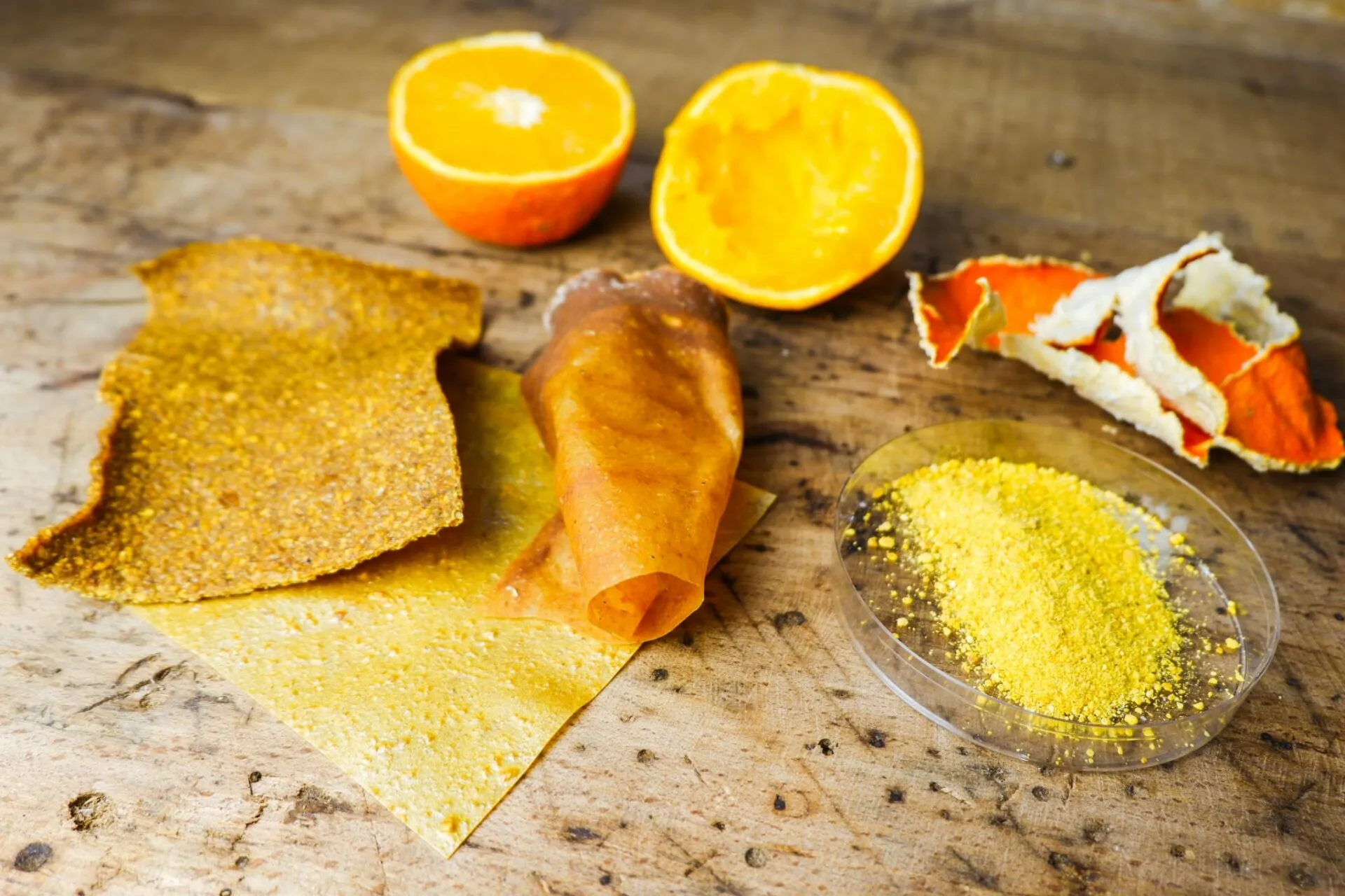 Halved oranges, orange peel and sheets of dried fruit leather on a rough wooden table, with a dish of yellow powder