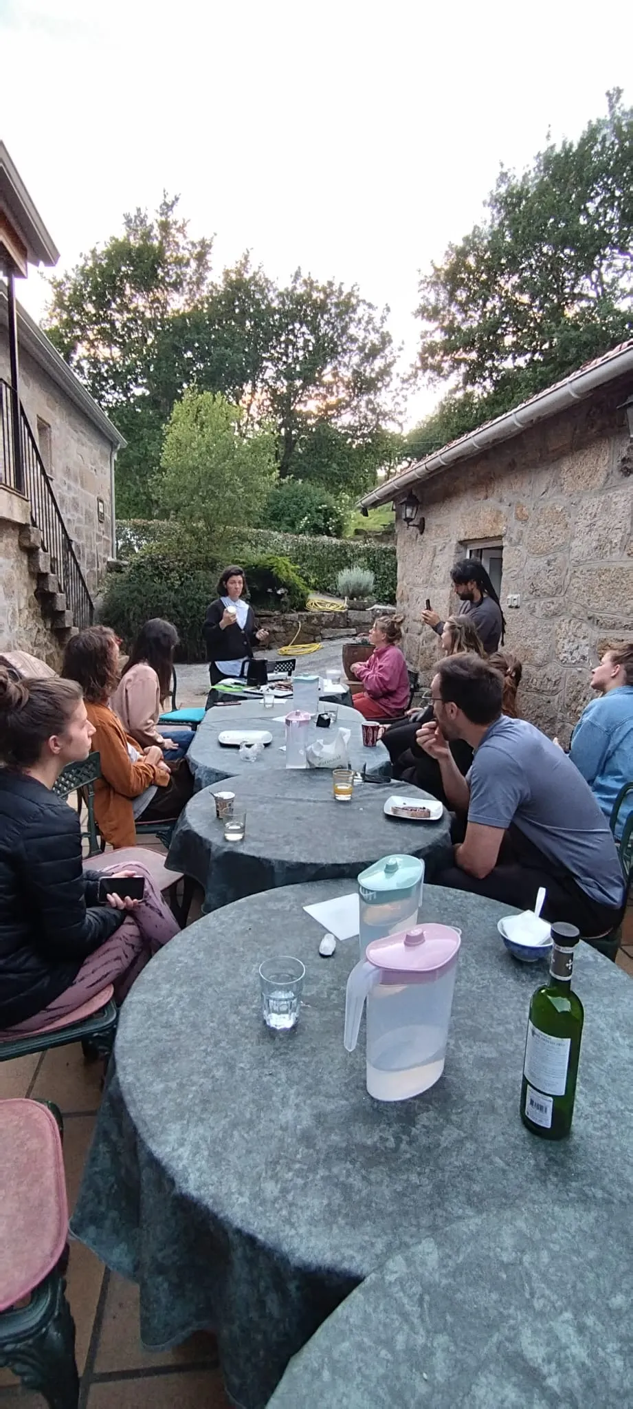 A woman speaking to a group seated around round tables on a stone terrace at dusk