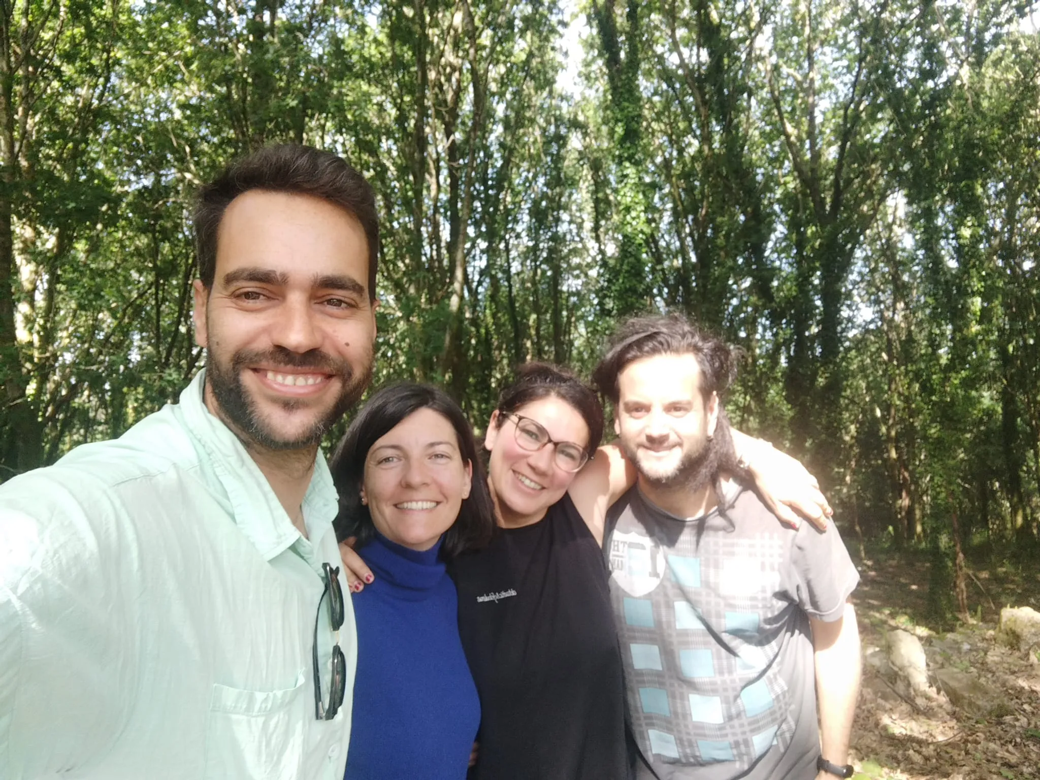 Selfie of four smiling people with arms around each other in a sunlit green wood