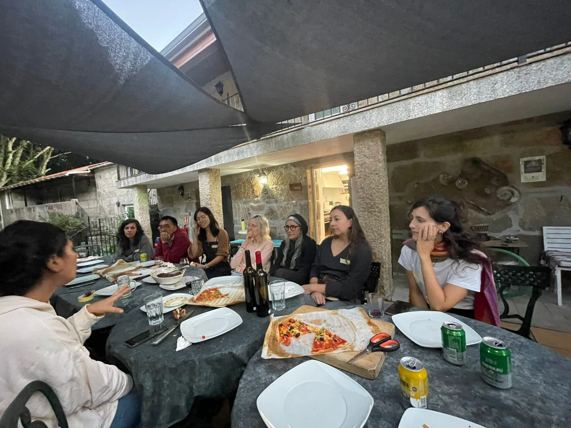 Group of people sitting around a table sharing pizza and wine at a community dinner at Anceu Coliving