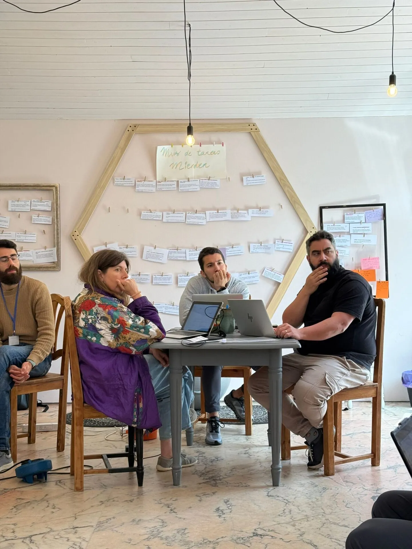 Four people with laptops at a small table in front of a wall of paper cards pegged inside a wooden hexagon labelled ‘Muro de tareas’