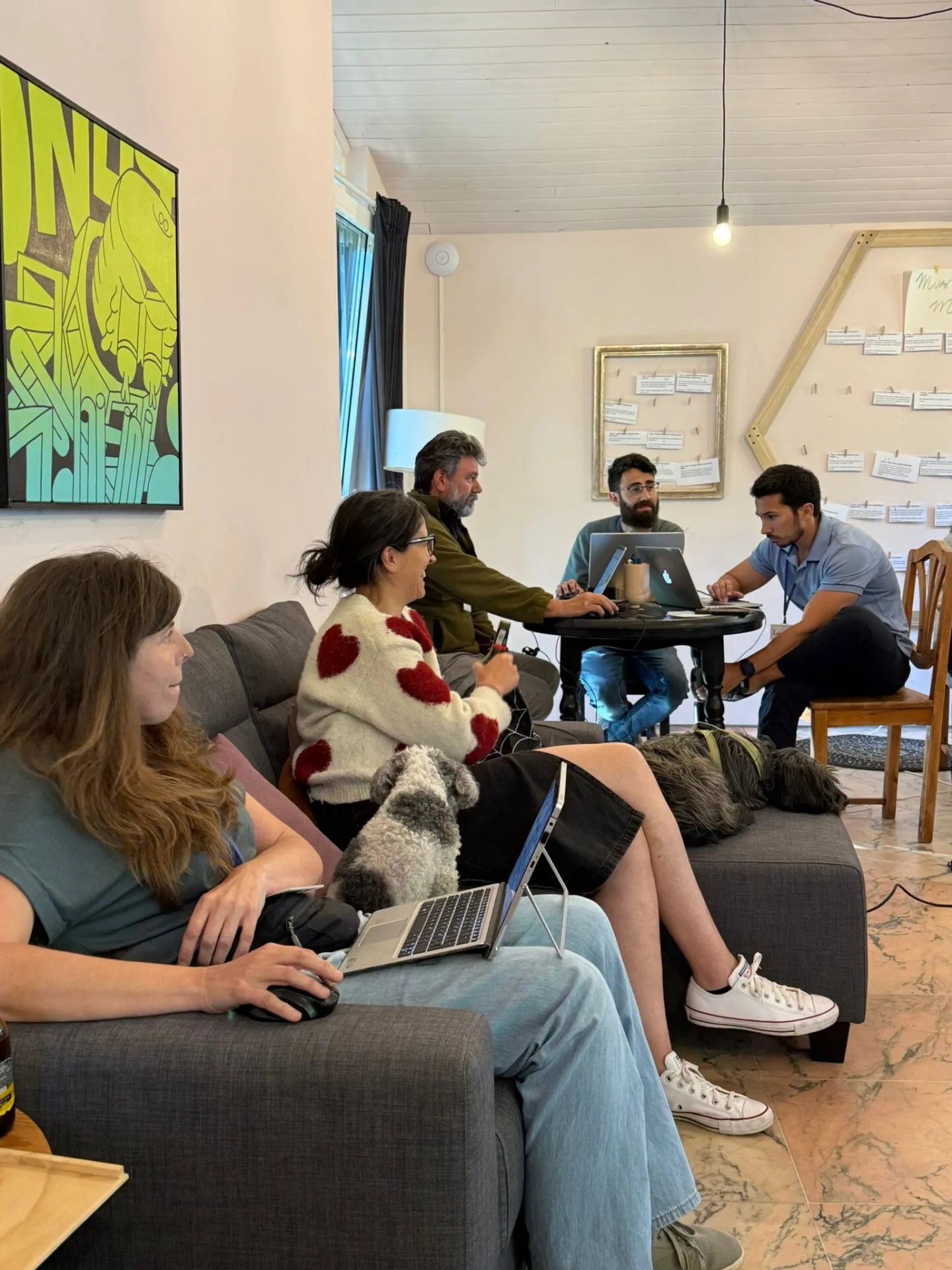 Six people working on laptops on sofas and a round table, with two dogs resting beside them