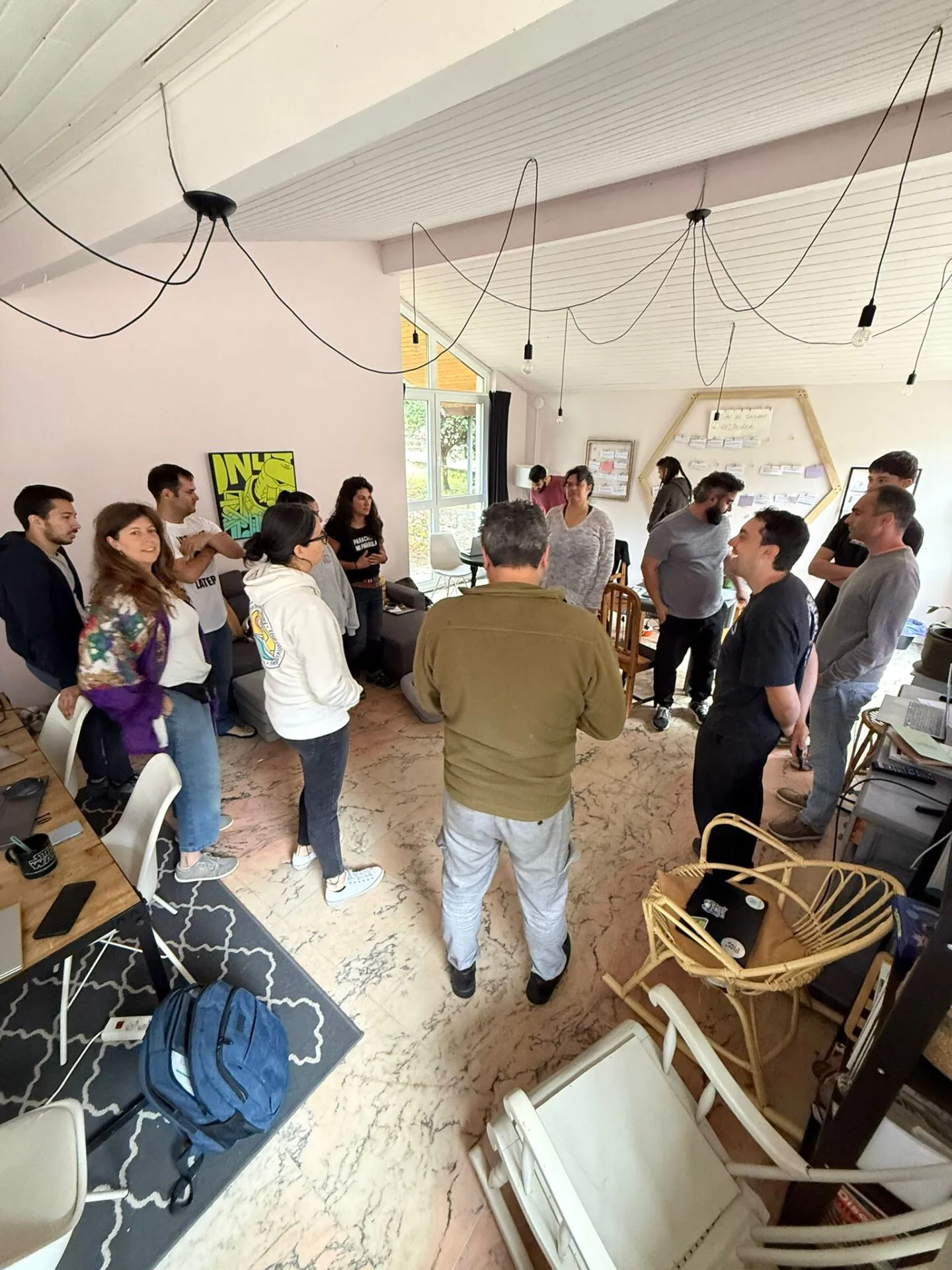 About fifteen people standing in a circle talking in the coworking room, laptops on a wooden table nearby