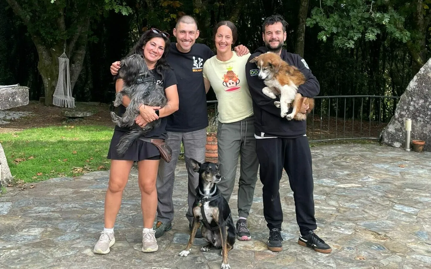 Four smiling people posing arm in arm on a stone terrace, two of them holding small dogs and a black dog at their feet