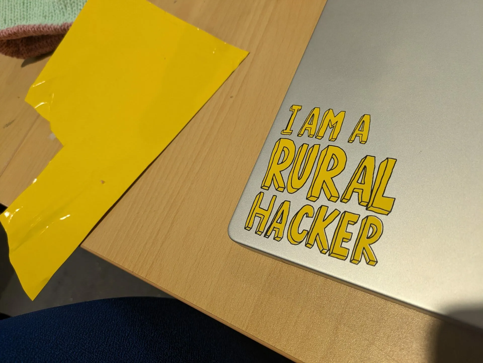 A laptop lid with a yellow sticker reading I AM A RURAL HACKER, lying on a wooden table beside a piece of yellow tape