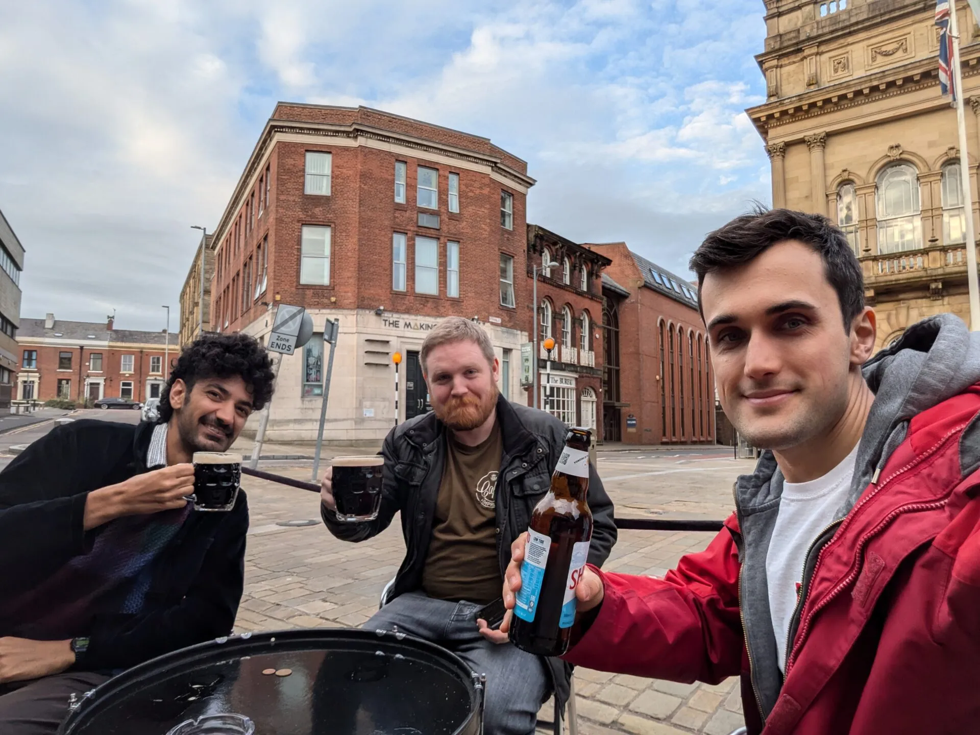 Three men raising beers at an outdoor table on a British city street, red brick buildings and a Union flag behind