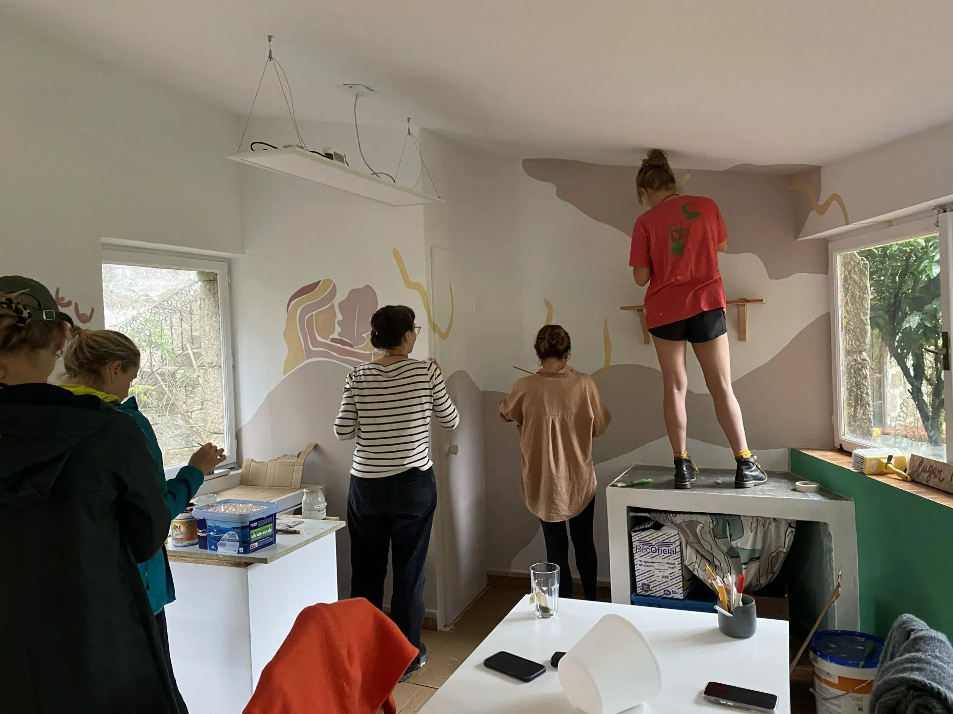 Four people painting the atelier walls in dusty pink and ochre, one standing on the worktop to reach the ceiling line