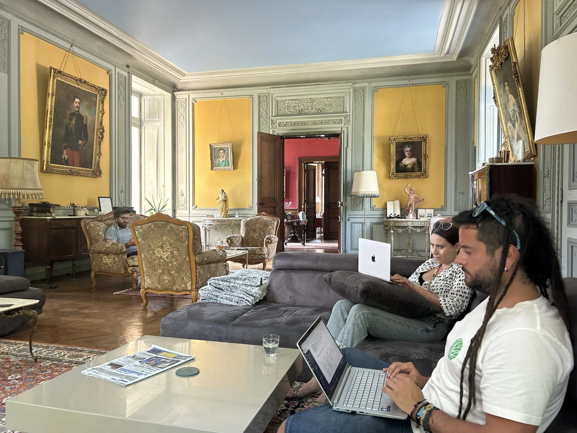 Three people working on laptops in a château salon with yellow panelled walls and gilt-framed portraits