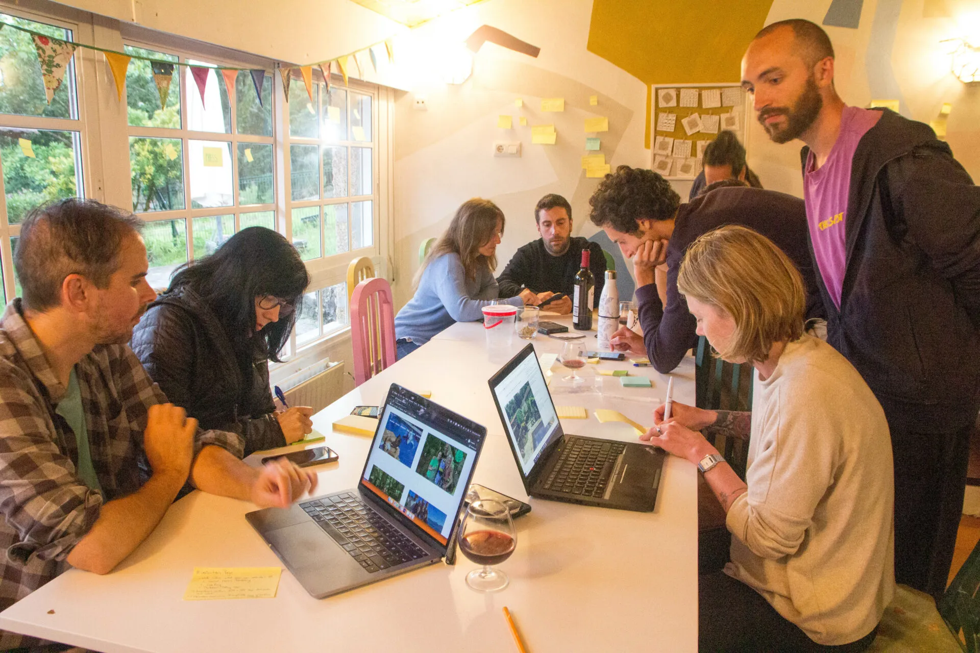 Seven people with laptops and sticky notes around a white table in a bright room with bunting and a large window