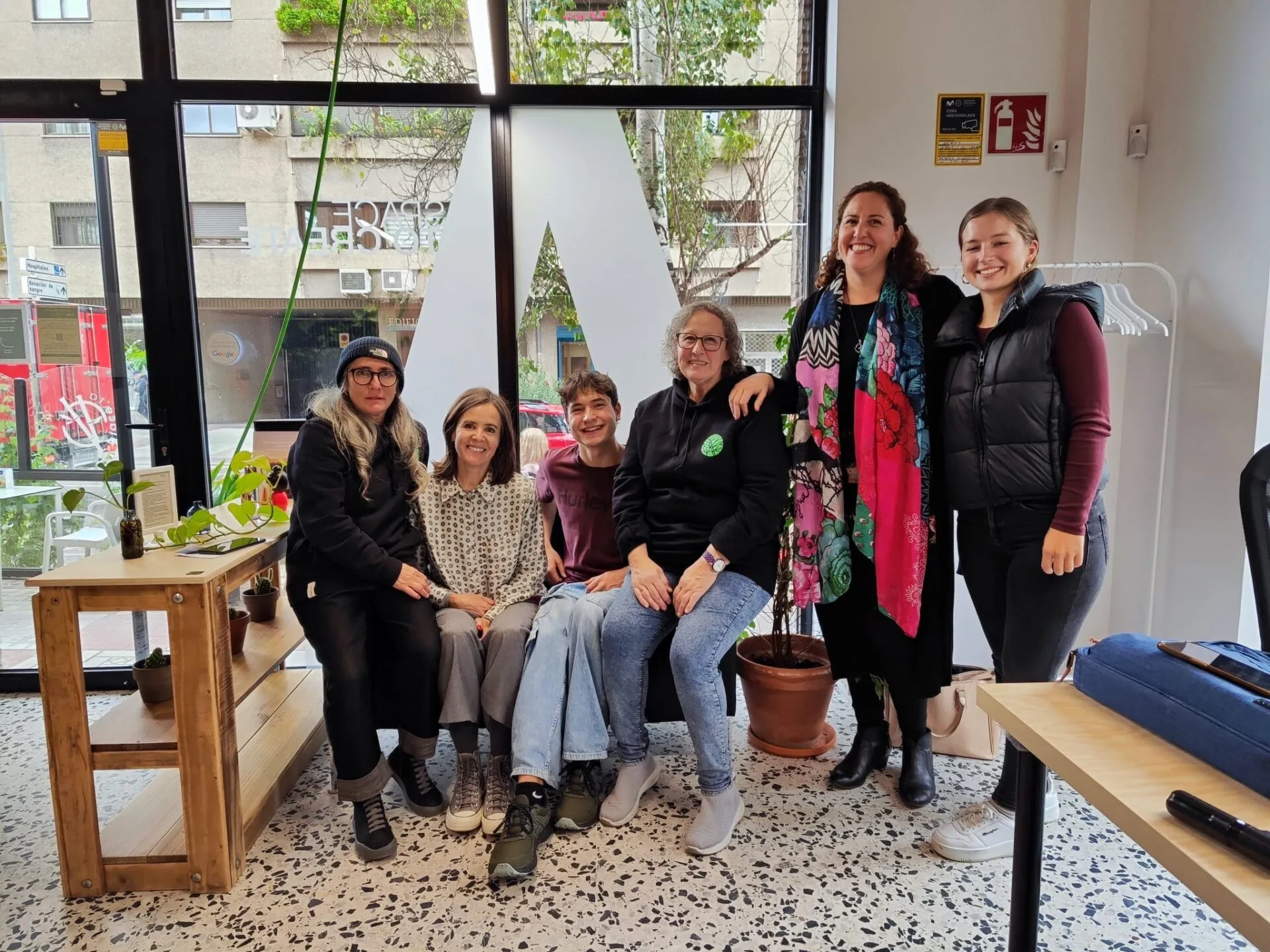 Six people posing together in a bright street-level office with a large logo on the glass front and a terrazzo floor