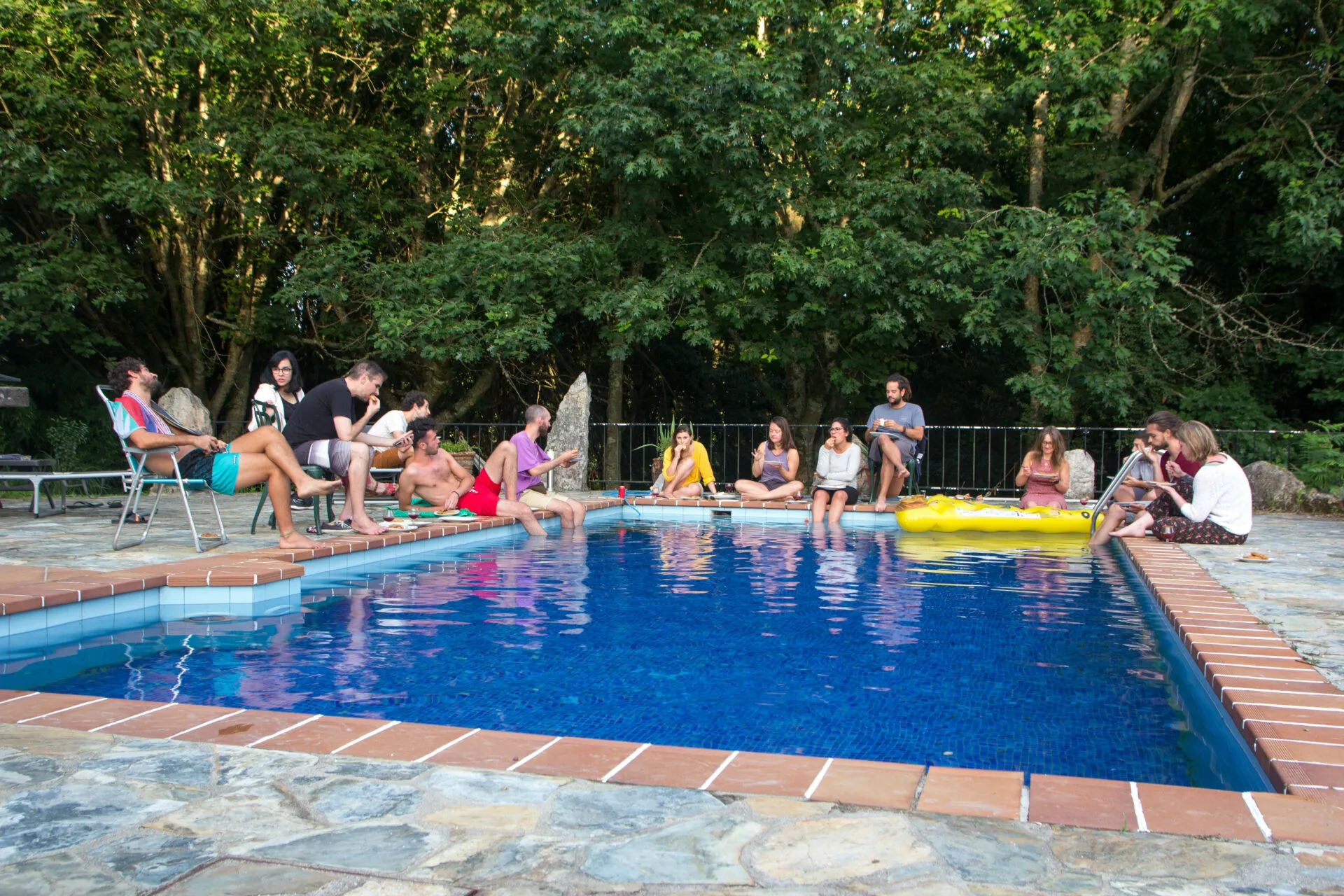 People lounging on chairs and loungers around a blue swimming pool in the evening, ringed by trees