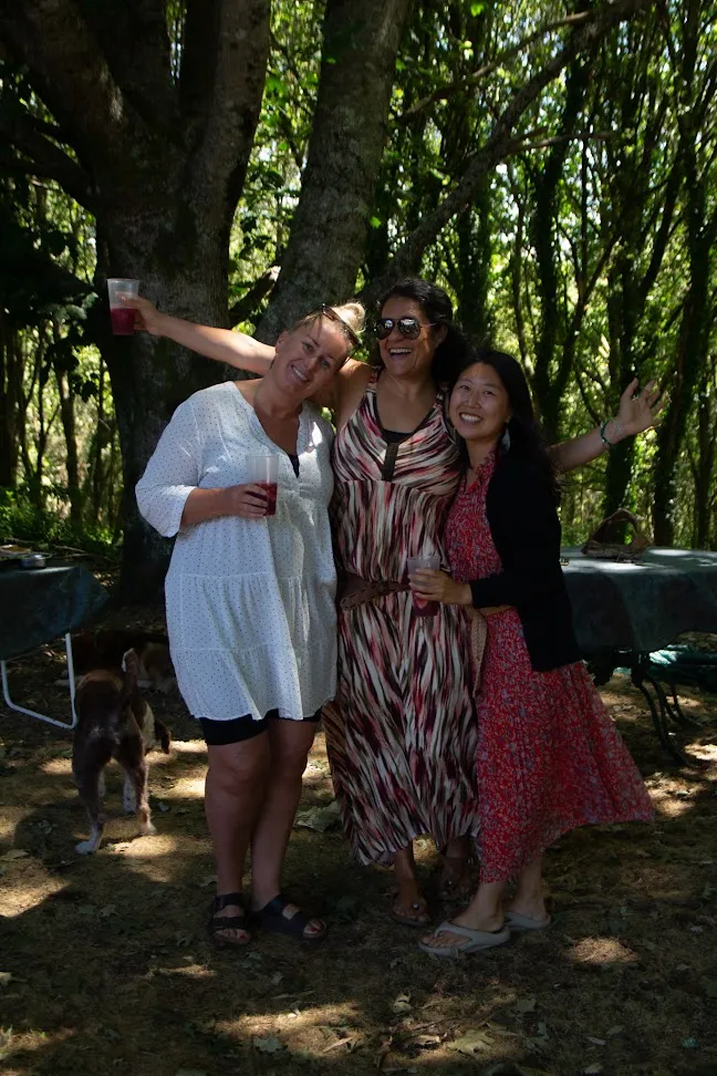 Three women laughing arm in arm with drinks in hand under shady trees, a dog wandering behind them